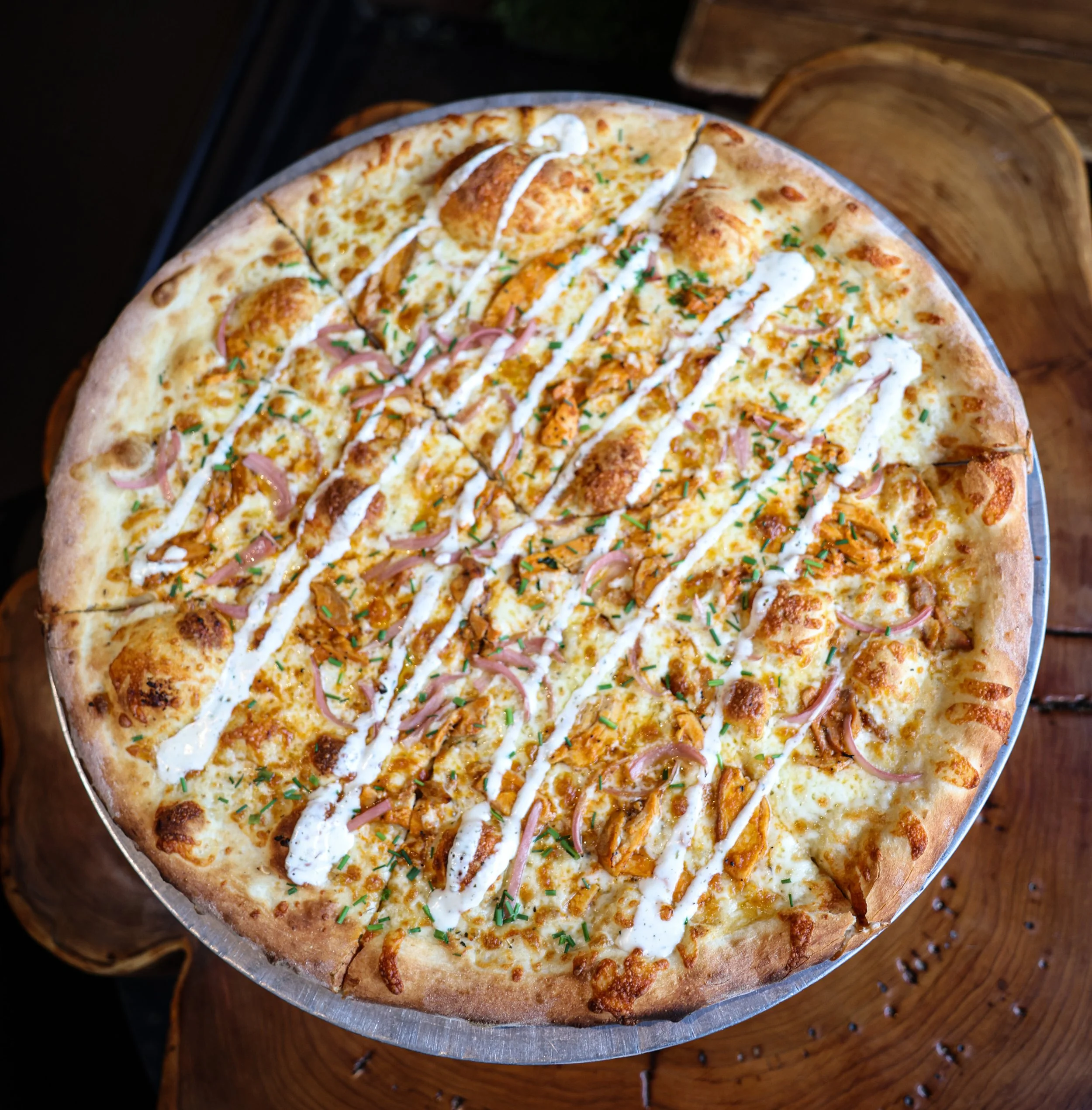 A pizza with cheese, sliced onions, shredded chicken, and green herbs drizzled with ranch or white sauce, served on a metal tray on a wooden table.