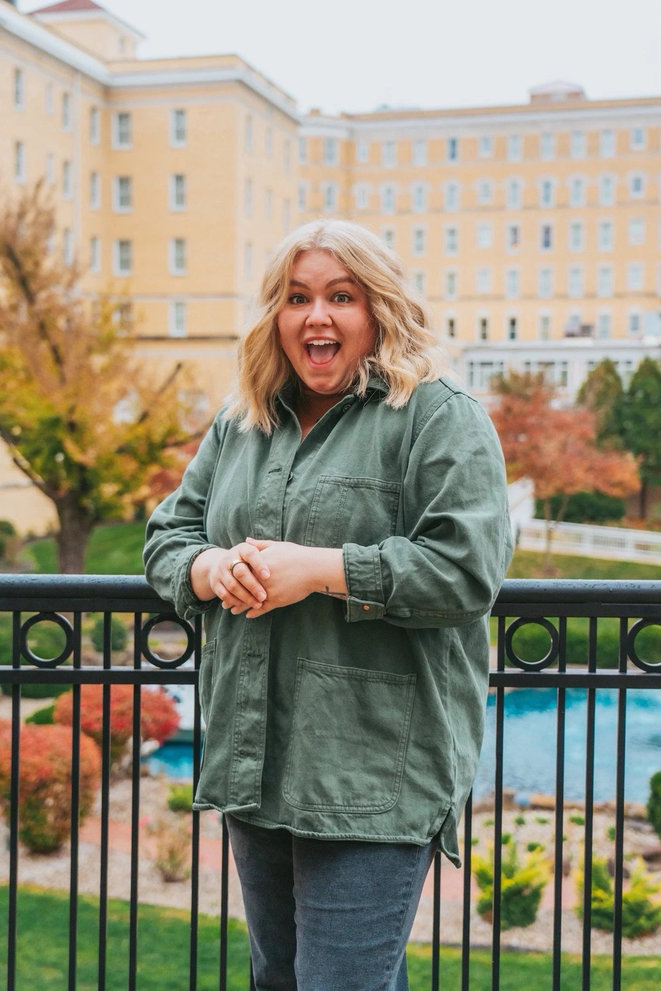 Blonde woman with excited facial expression. There is a pond and nature background with yellow brick building.