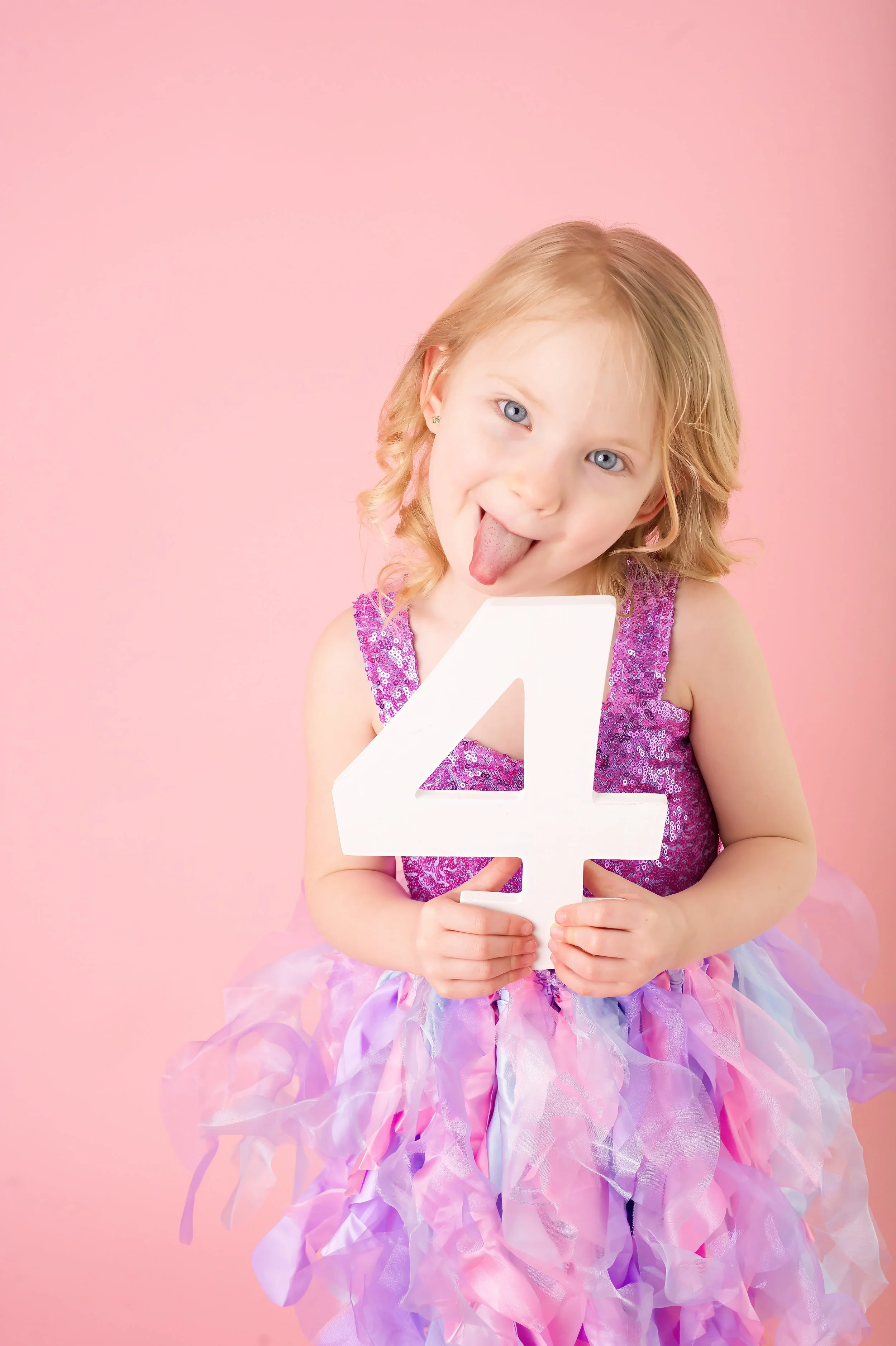 A young girl with blonde hair and blue eyes sticking out her tongue, holding a large white number 4, wearing a purple sparkling top and a pastel-colored tutu skirt, against a pink background.