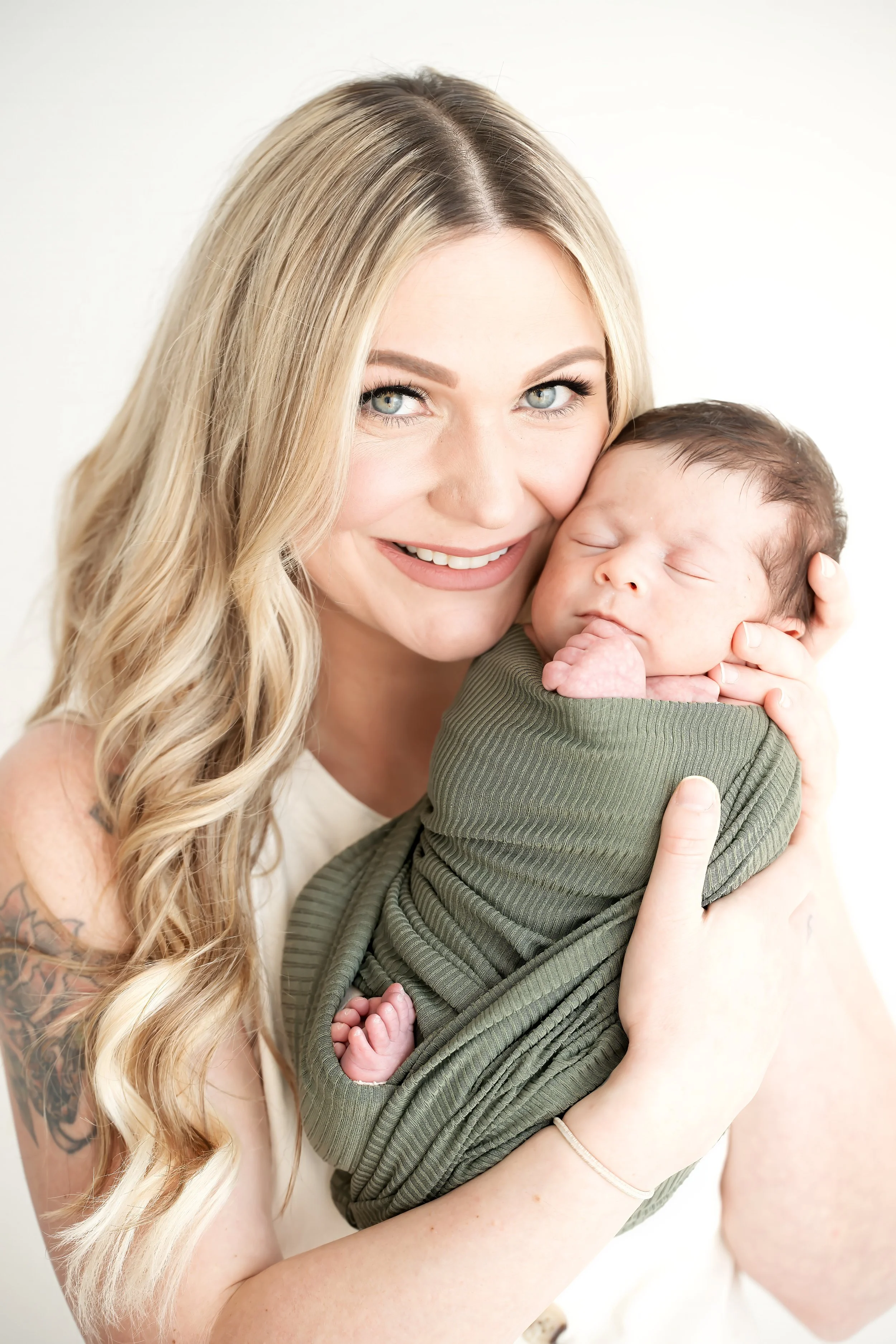 A woman with long blonde hair and tattoos on her arm is holding a sleeping baby wrapped in a green blanket.