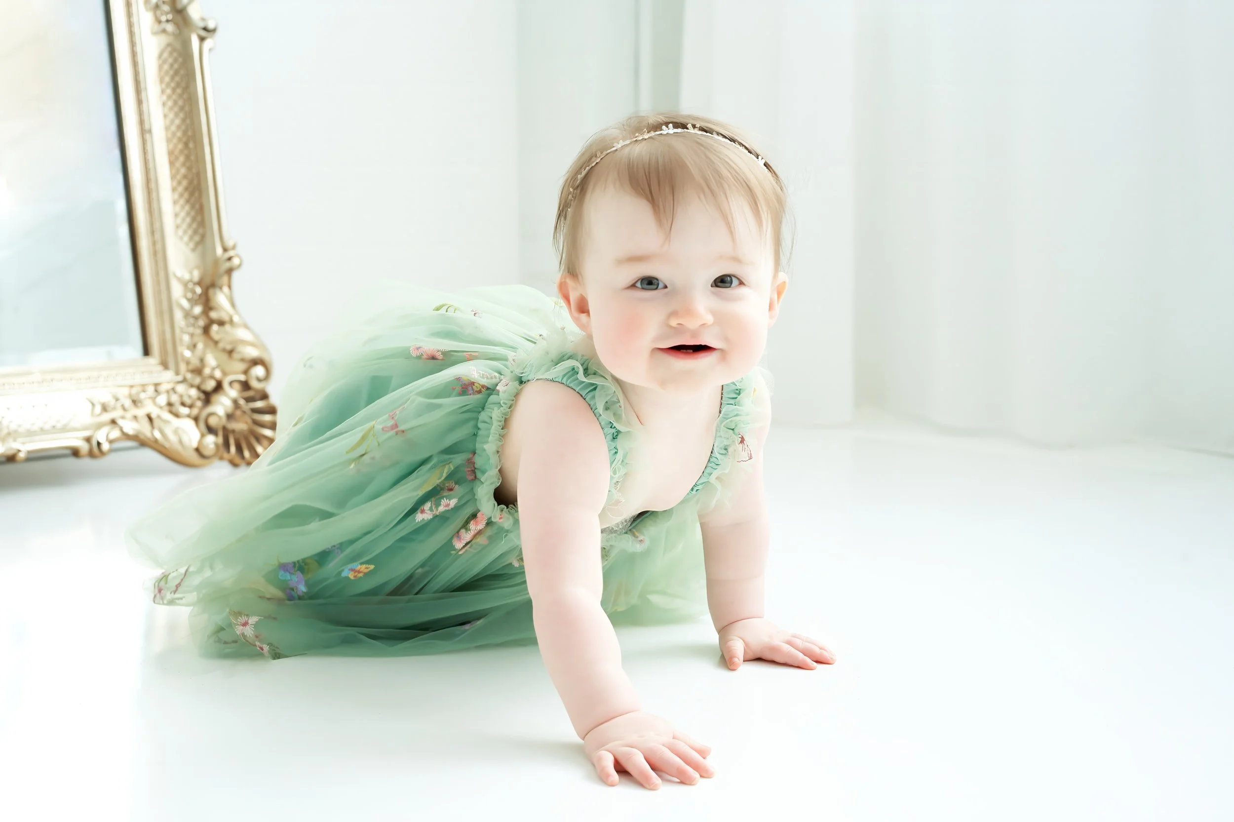 A smiling toddler girl with light brown hair in a thin white headband, crawling on a white floor, wearing a green dress with colorful embroidery.