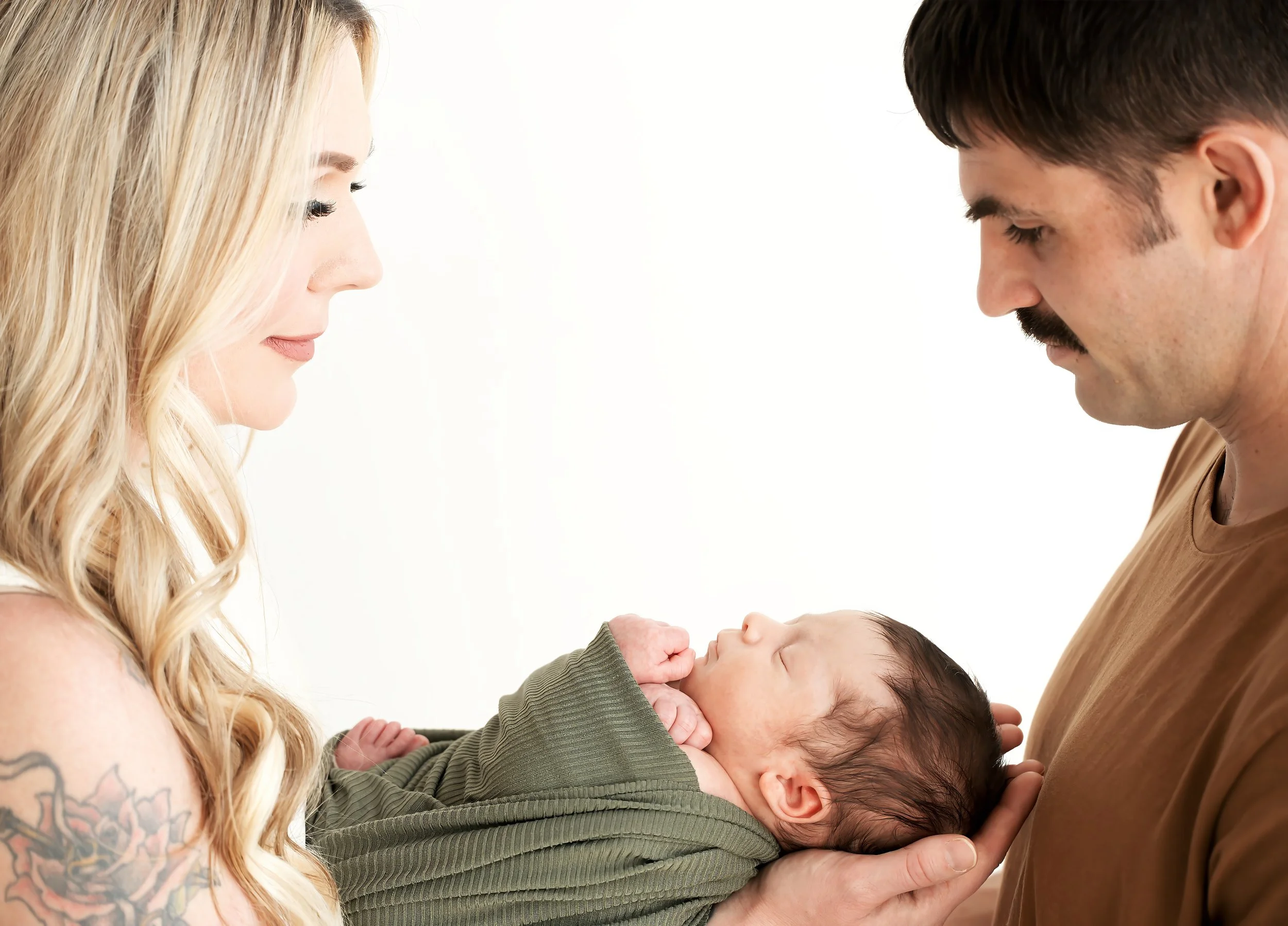 A blonde woman and a man holding a newborn baby, facing each other with eyes closed, in a bright setting.