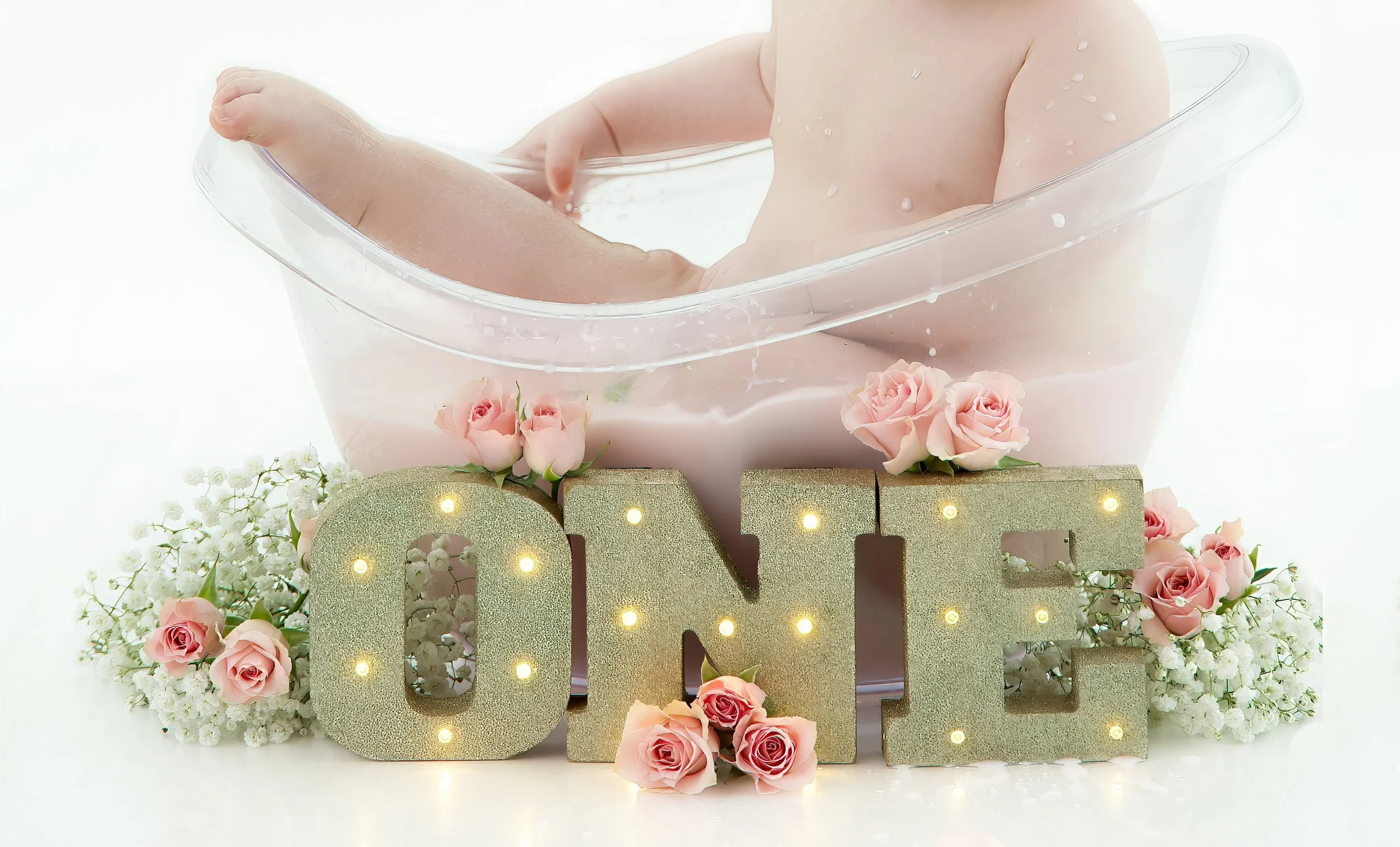 Baby in a clear bathtub with water droplets, surrounded by pink roses, white flowers, and gold decorative letters spelling 'ONE' with lights inside.