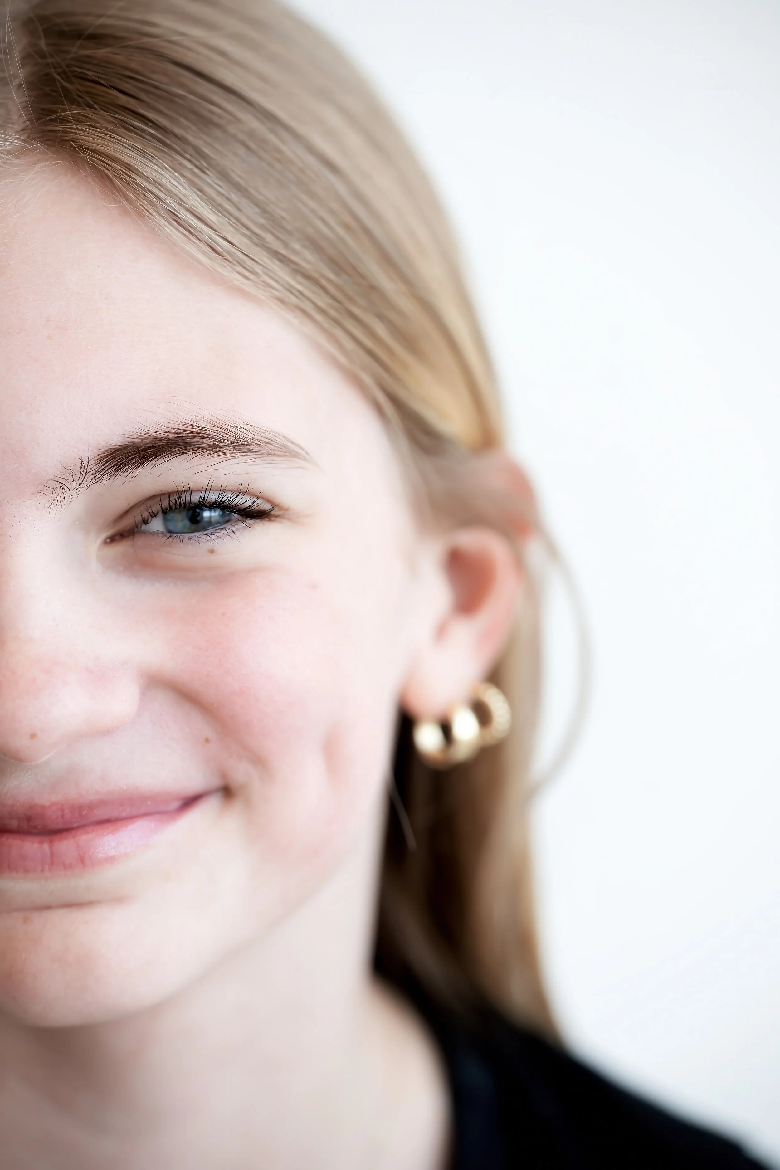 Close-up of a smiling woman with blonde hair, blue eyes, and gold earrings, showing part of her face from the nose up.