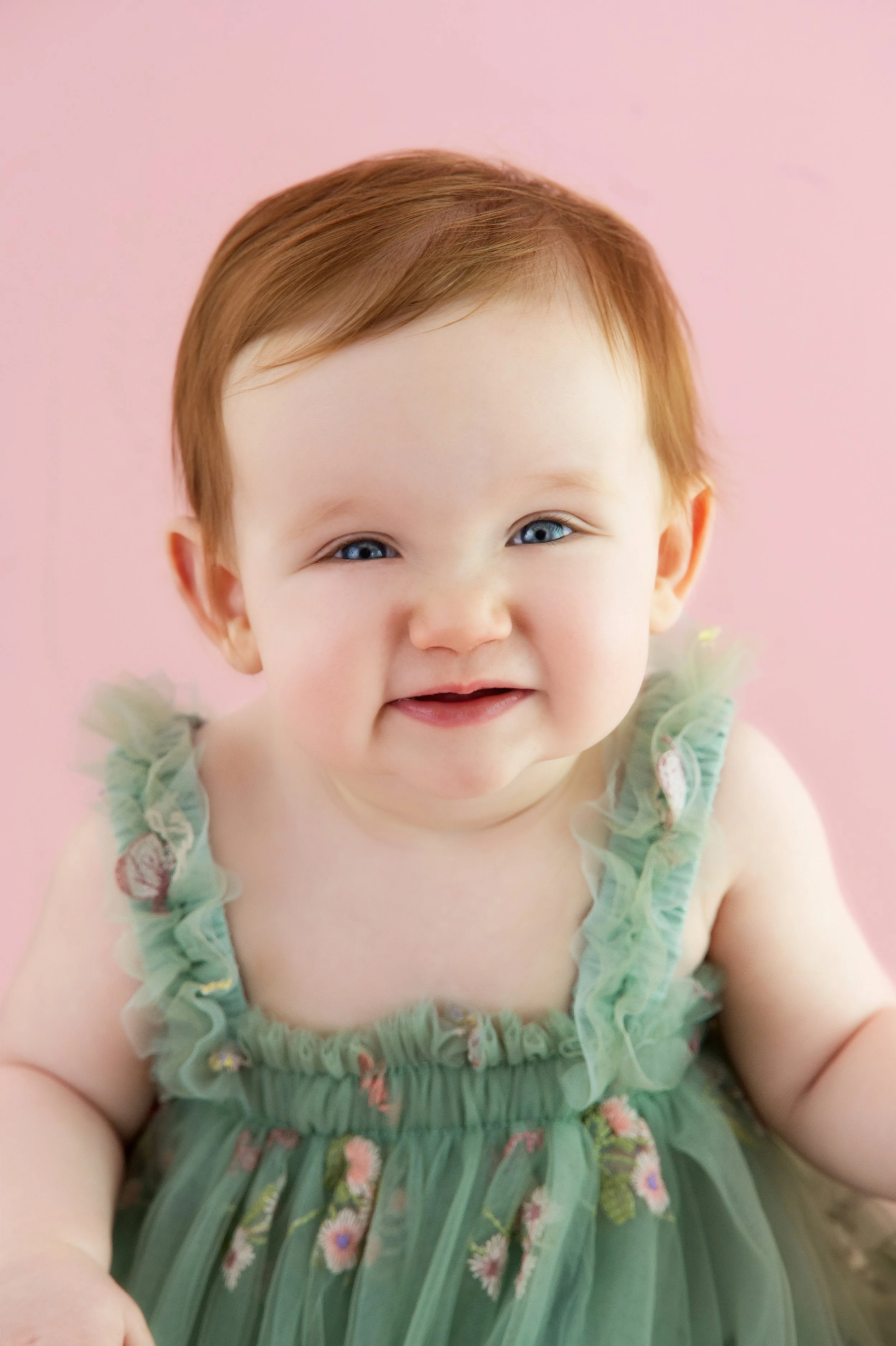 Close-up of a smiling baby girl with blue eyes, wearing a green dress with pink floral embroidery, against a pink background.