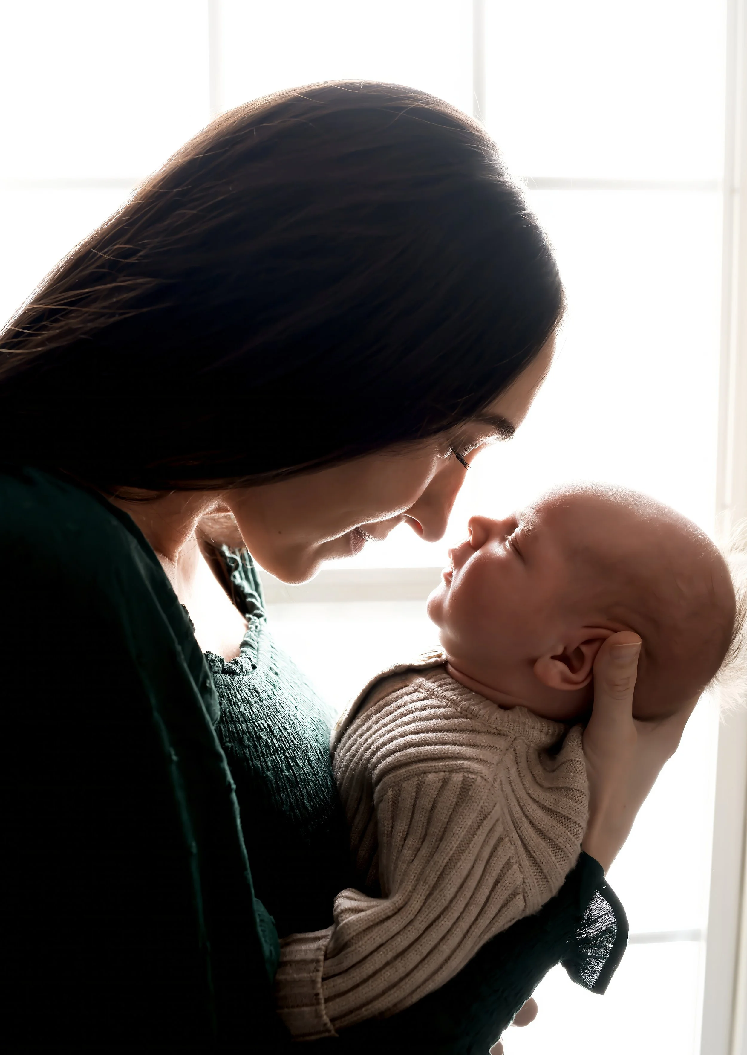 A woman with dark hair holding a baby, close to each other with their foreheads touching, near a bright window.