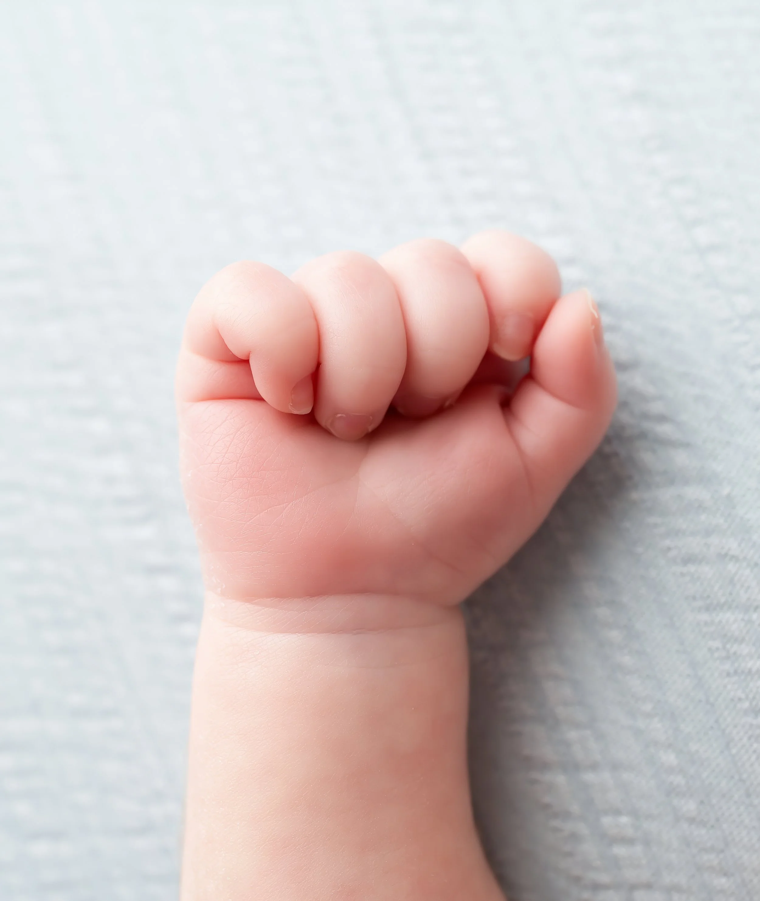 Close-up of a newborn baby's fist with tiny fingers clenched, resting on a soft, light-colored fabric surface.