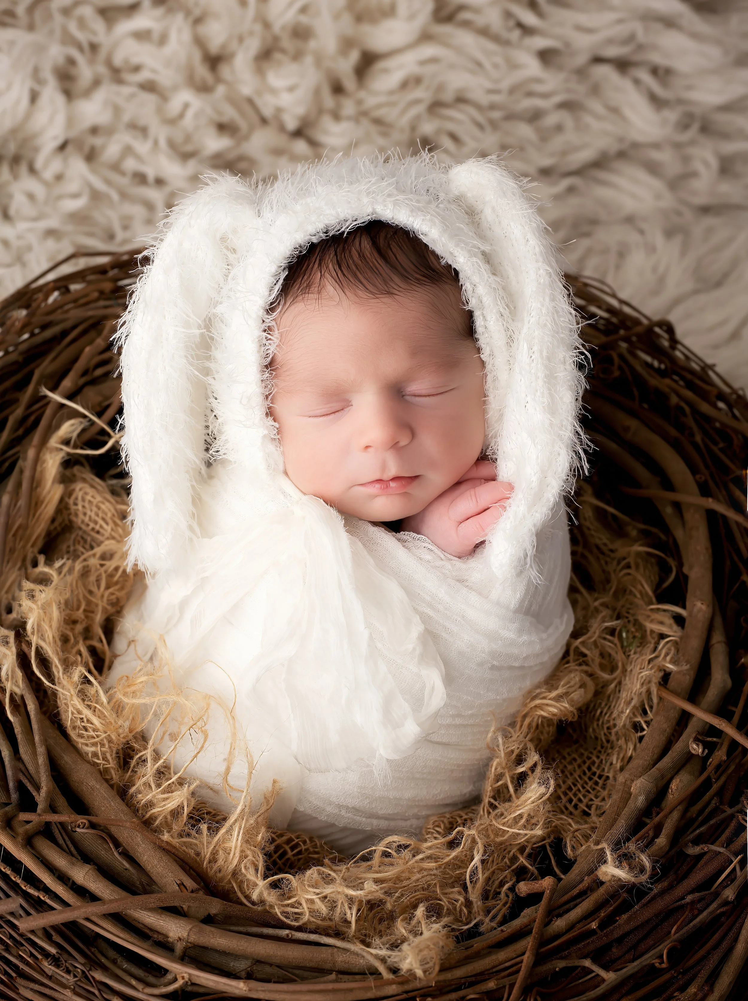 Close-up of a sleeping baby in a woven basket, wearing a white hooded garment, surrounded by soft blankets.
