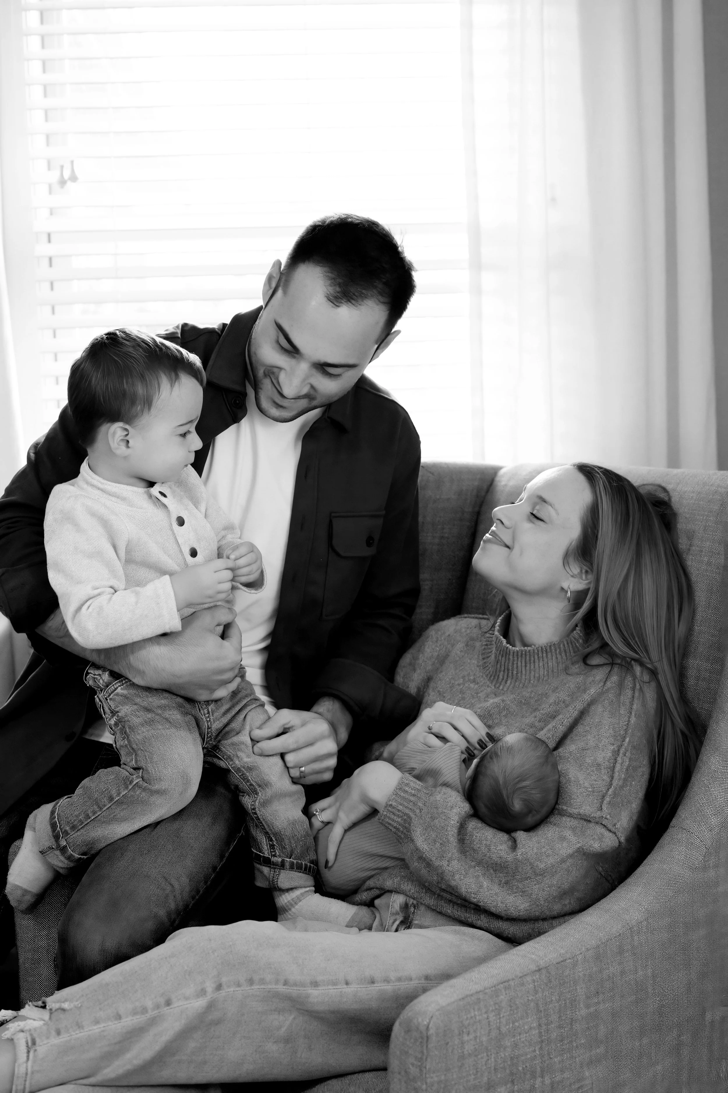 A family of four on a sofa, with a man holding a young boy, a woman holding a baby, and another small child at the bottom. They are smiling and sharing a joyful moment together.