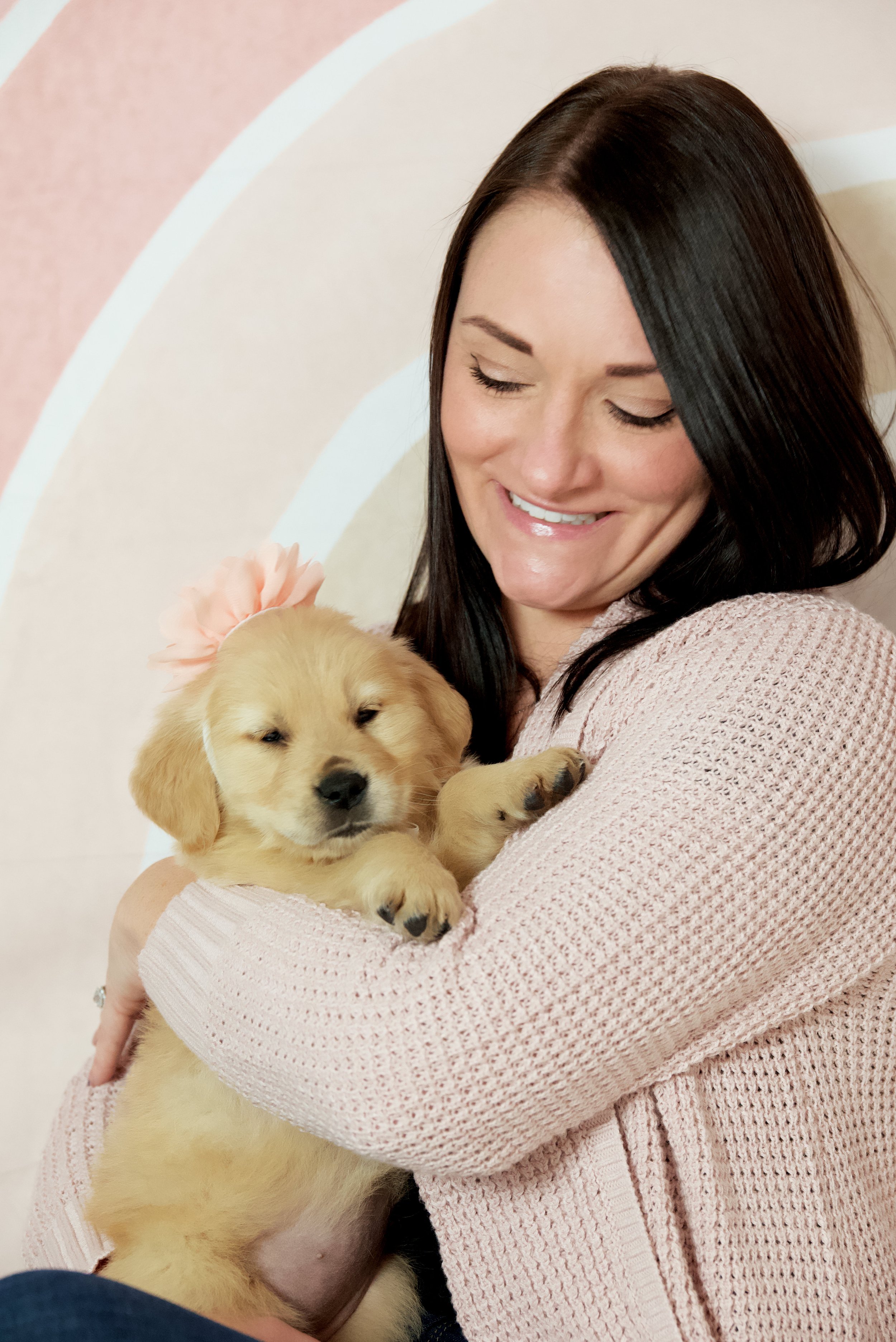 Woman holding a golden retriever puppy wearing a pink flower headband, against a patterned background.