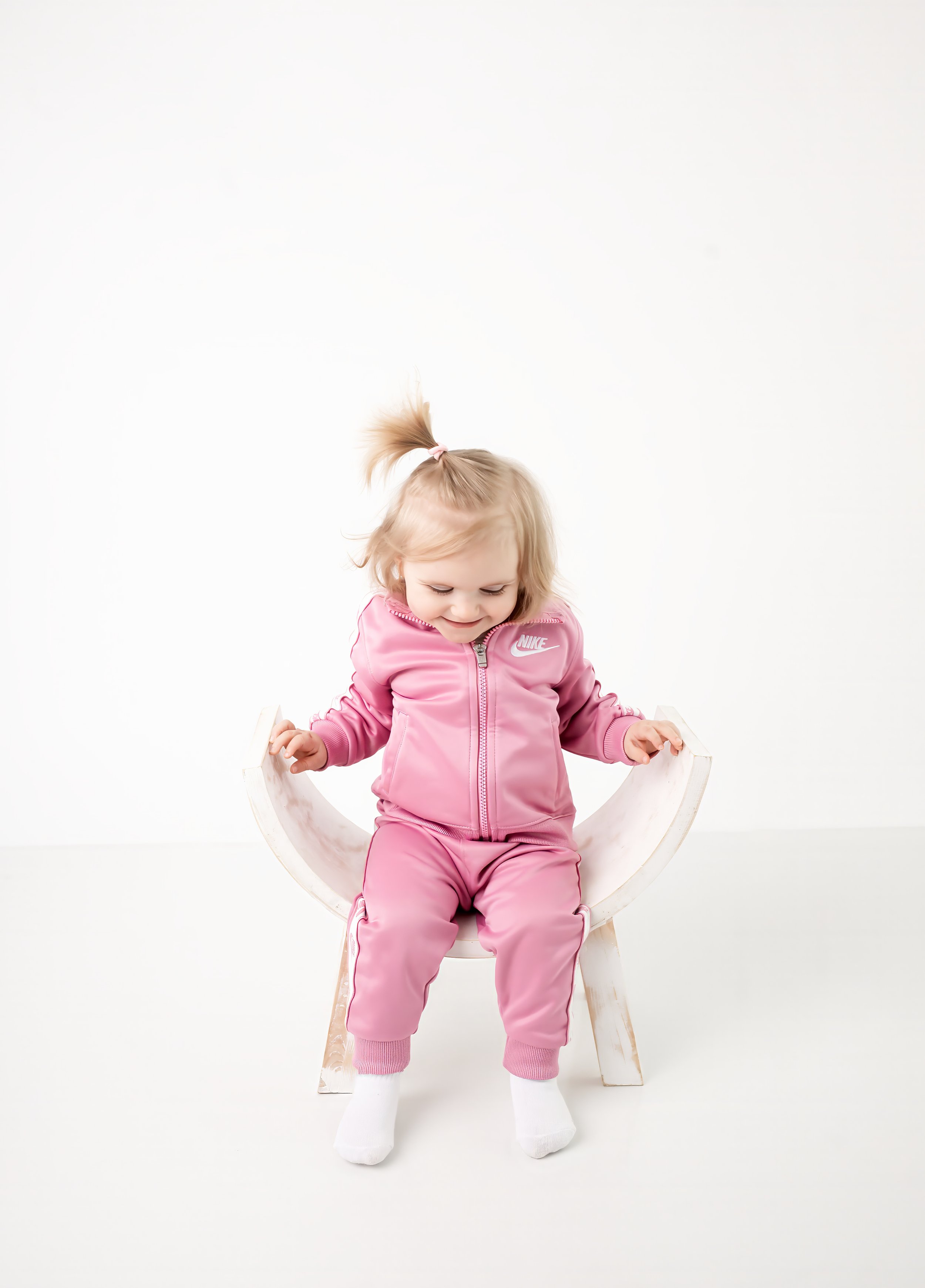 A young girl with blonde hair in a pink Nike tracksuit and white socks sitting on a wooden chair against a plain white background.