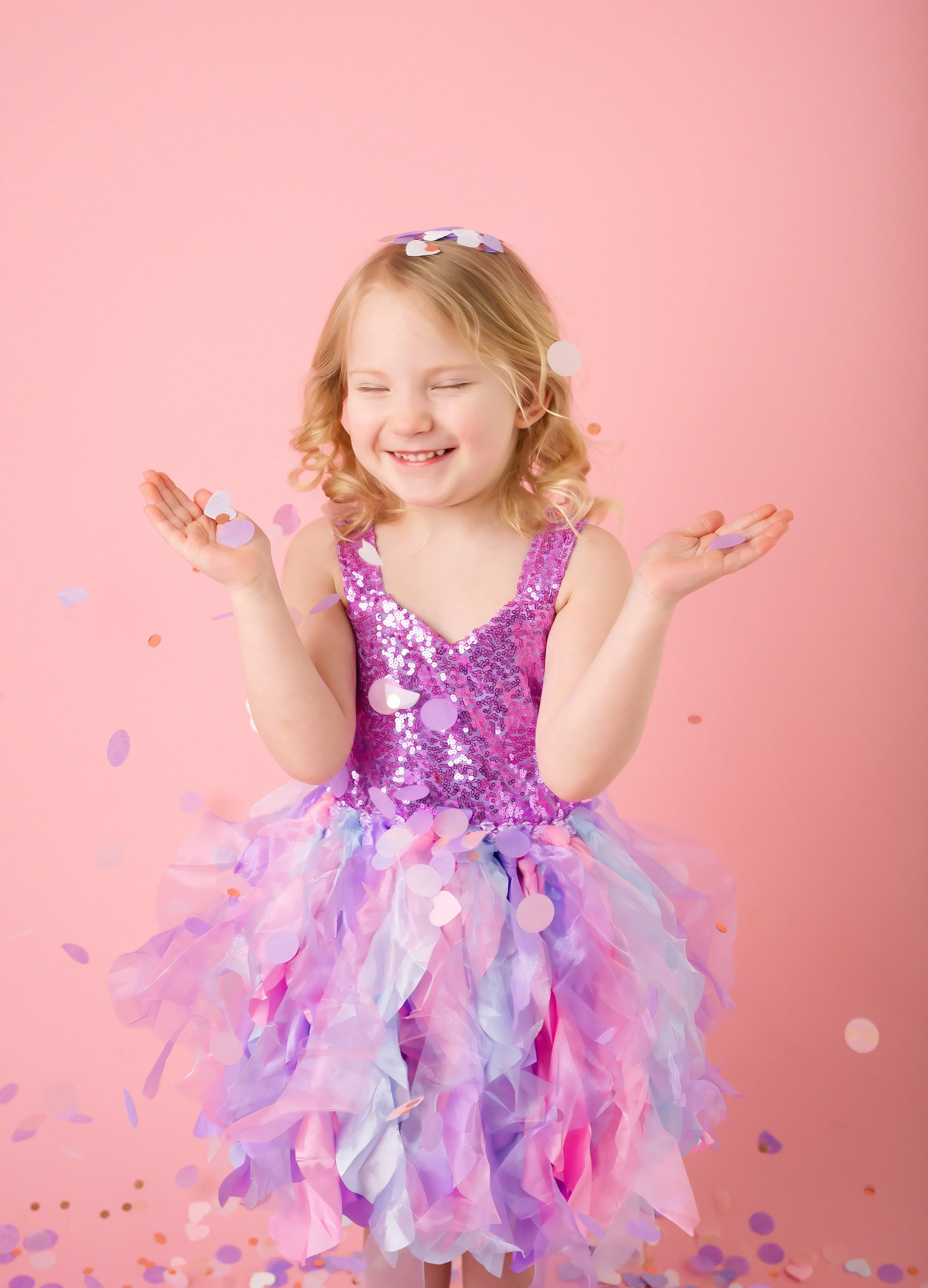 A young girl with curly blonde hair, dressed in a pink glittery top and layered pastel-colored tulle skirt, smiling with eyes closed as confetti falls around her against a pink background.