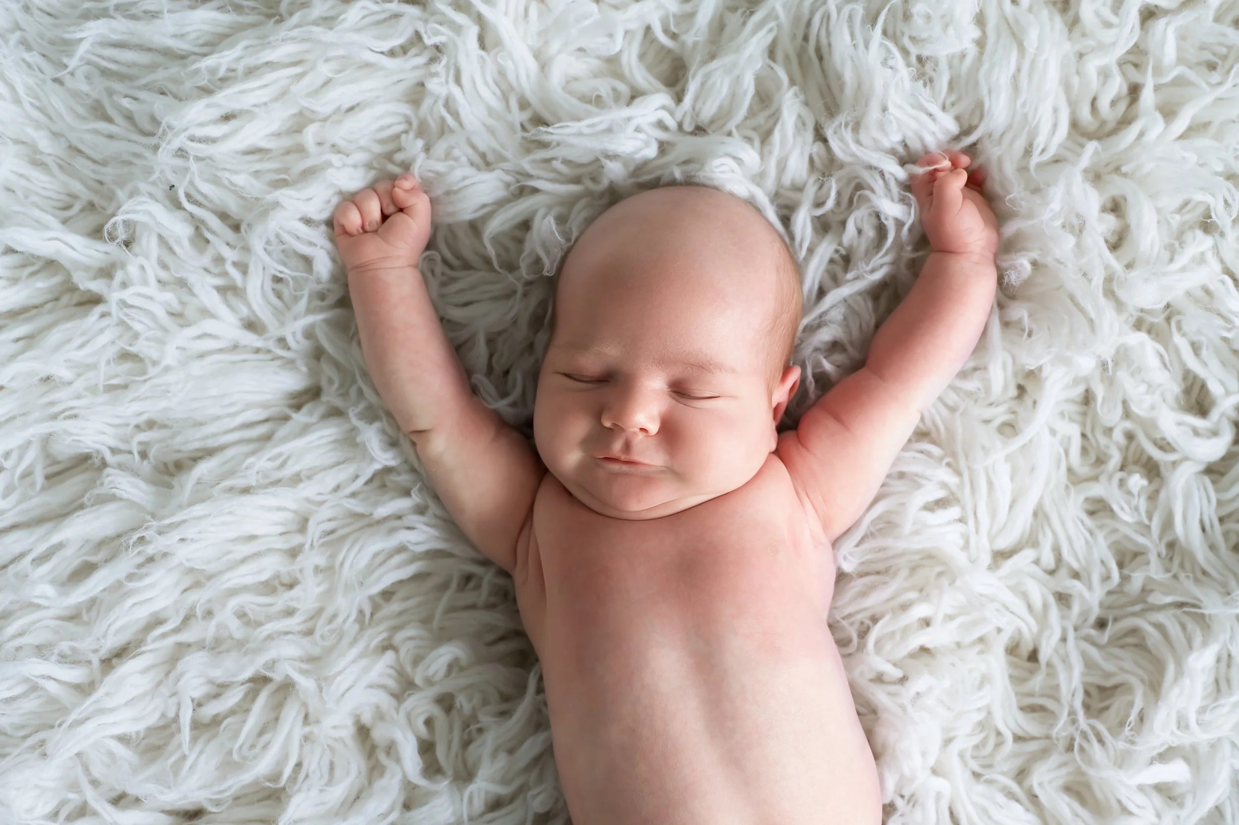 A newborn baby outstretched and smiling on a soft, fluffy white blanket..
