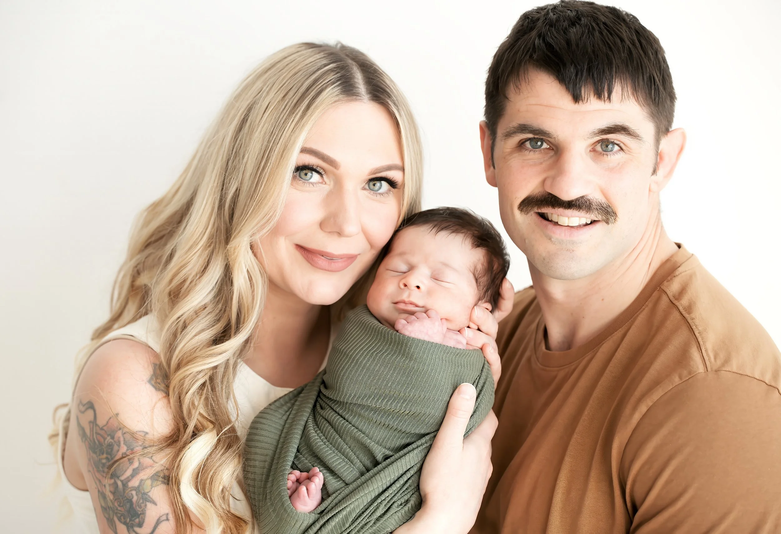 A happy family of three (mother, father, and newborn baby) smiling and posing against a white background.