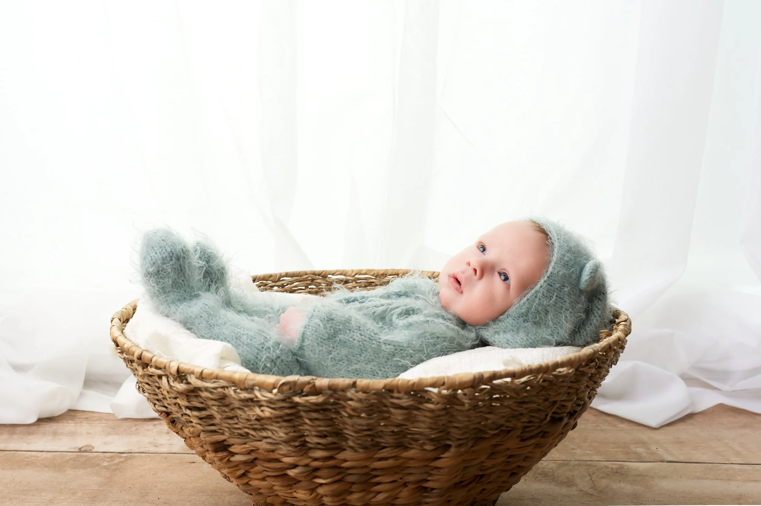 A baby lying in a wicker basket, wearing a soft gray hat and matching sweater, looking up with wide eyes, against a white curtain background.