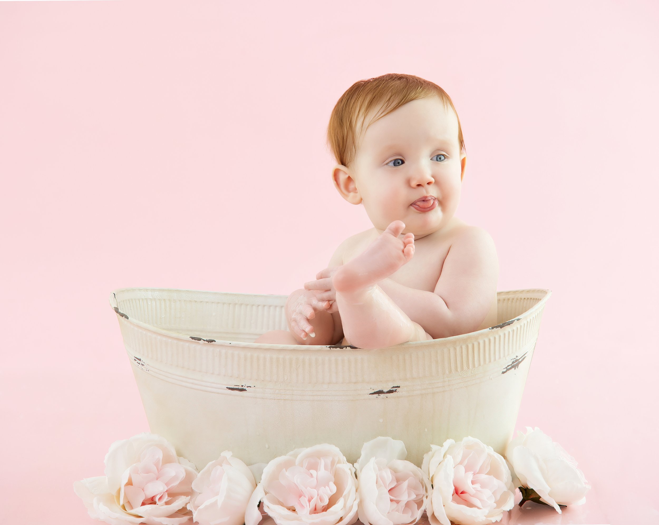 A young child with red hair and blue eyes sitting in a white bowl, sticking out tongue, surrounded by pink flowers on a pink background.