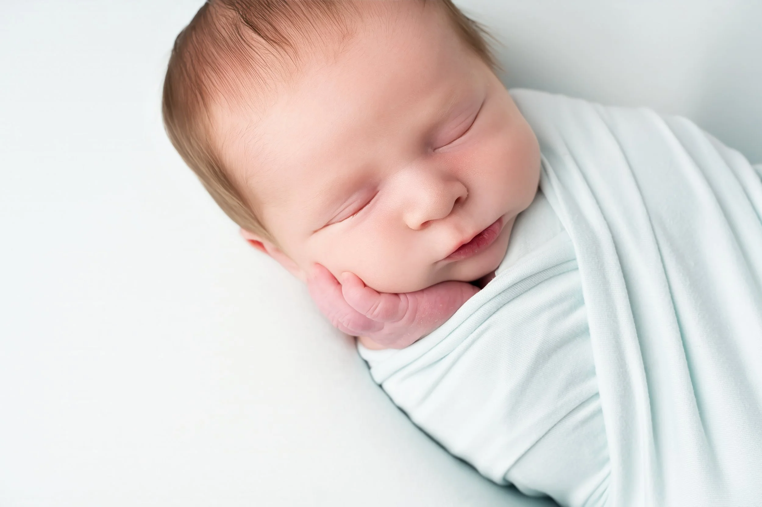 A close-up of a sleeping baby with light skin, closed eyes, and light brown hair, resting with hand near face, wearing a white outfit against a plain light background.