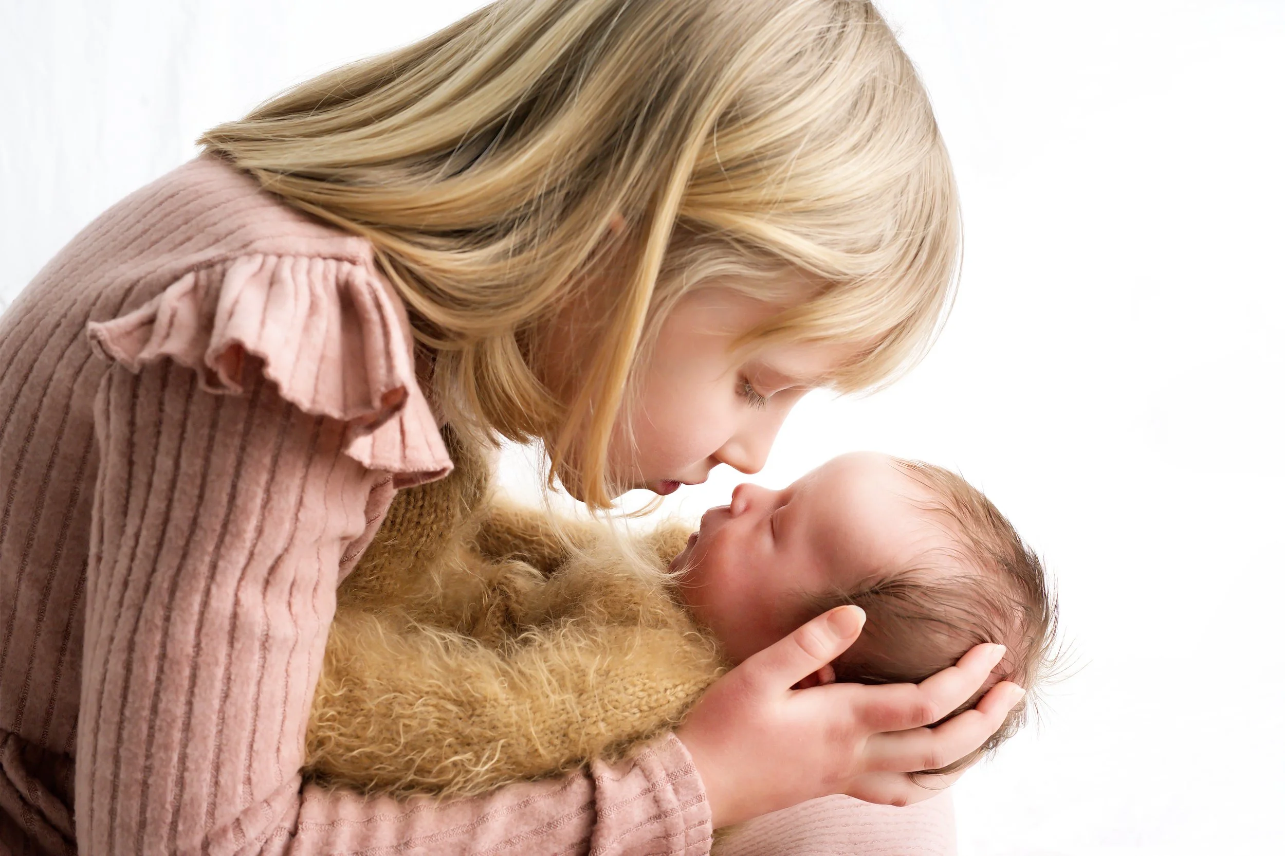 A woman with blonde hair and pink clothing gently holding and looking at a baby with similar hair texture. They are close in an intimate, caring pose against a white background.