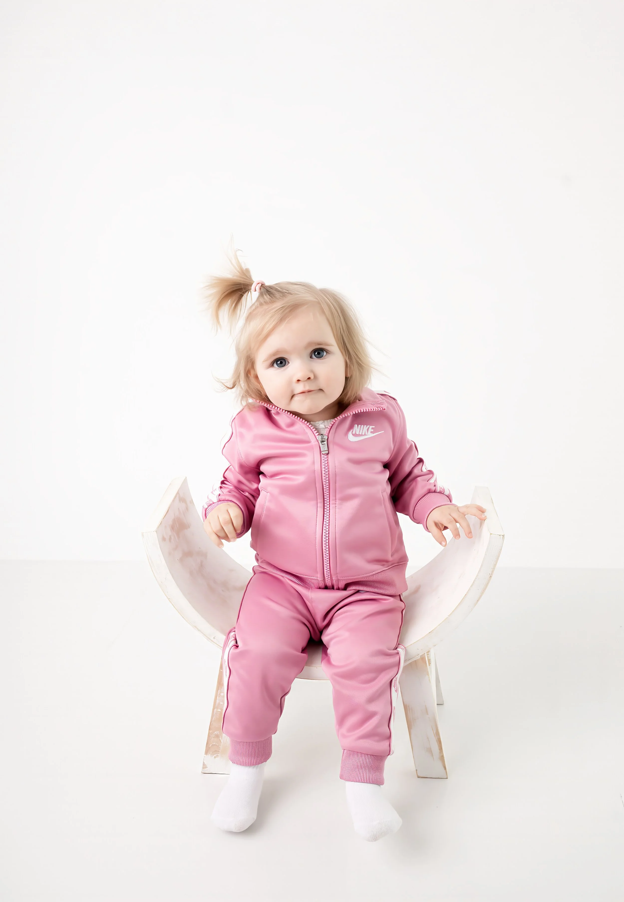 A young girl with blonde hair in a pink Nike tracksuit sitting on a white chair against a plain white background.