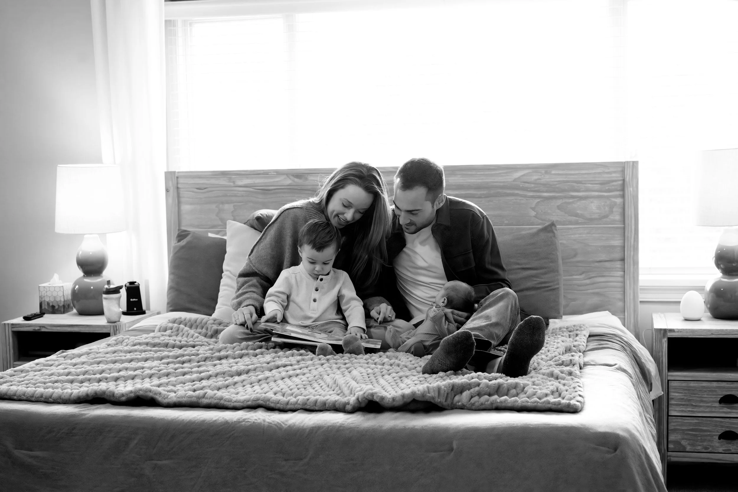 A family of four sitting on a bed in a bedroom, smiling and looking at a book together. The family consists of a woman, a man, a young boy, and a baby. The room is bright with large windows, curtains, and table lamps on nightstands.