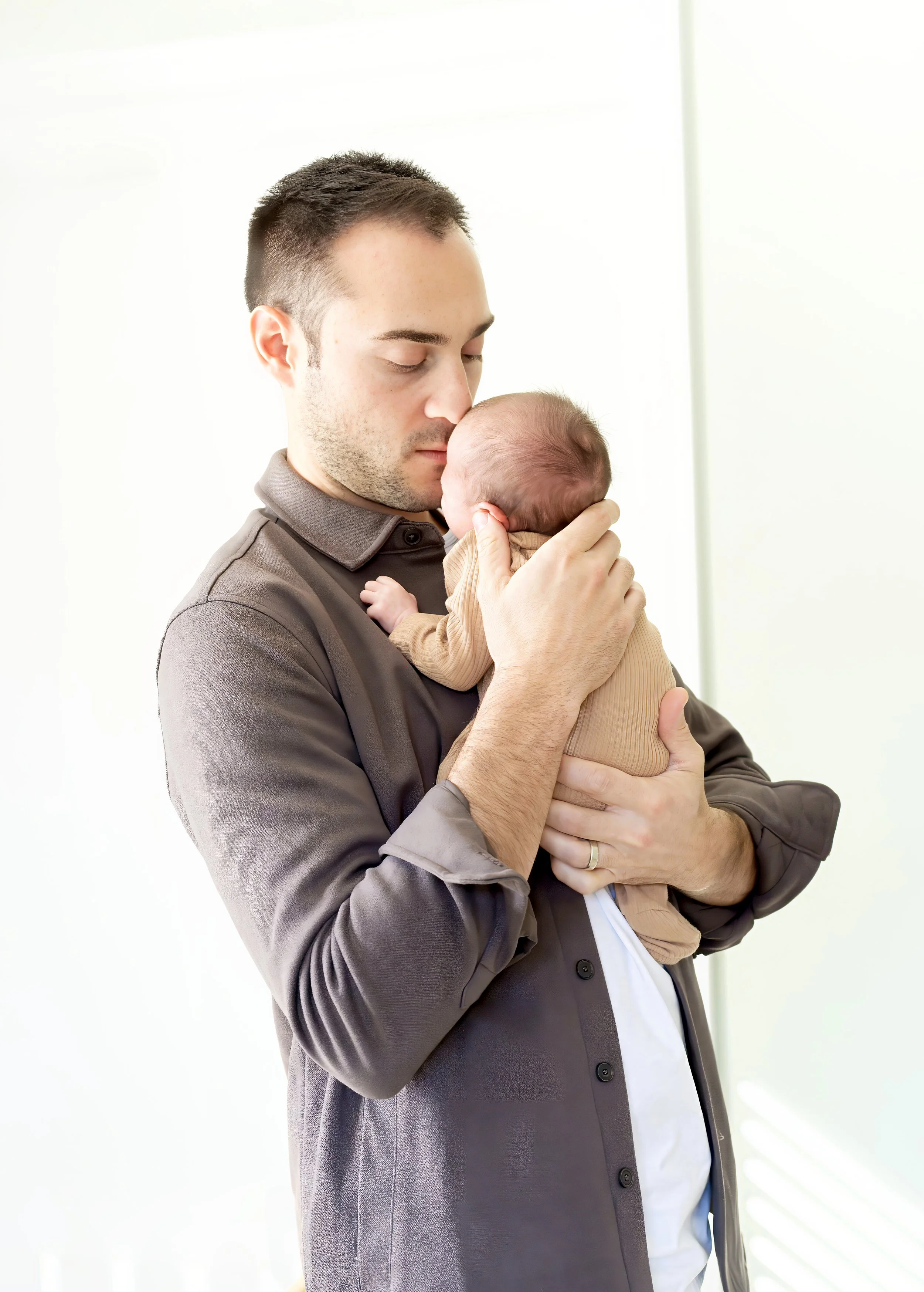 A man holding a baby close to his face, with both eyes closed, in a tender moment. The man is wearing a gray button-up shirt and the baby is dressed in a light brown outfit. The background is bright and blurred.
