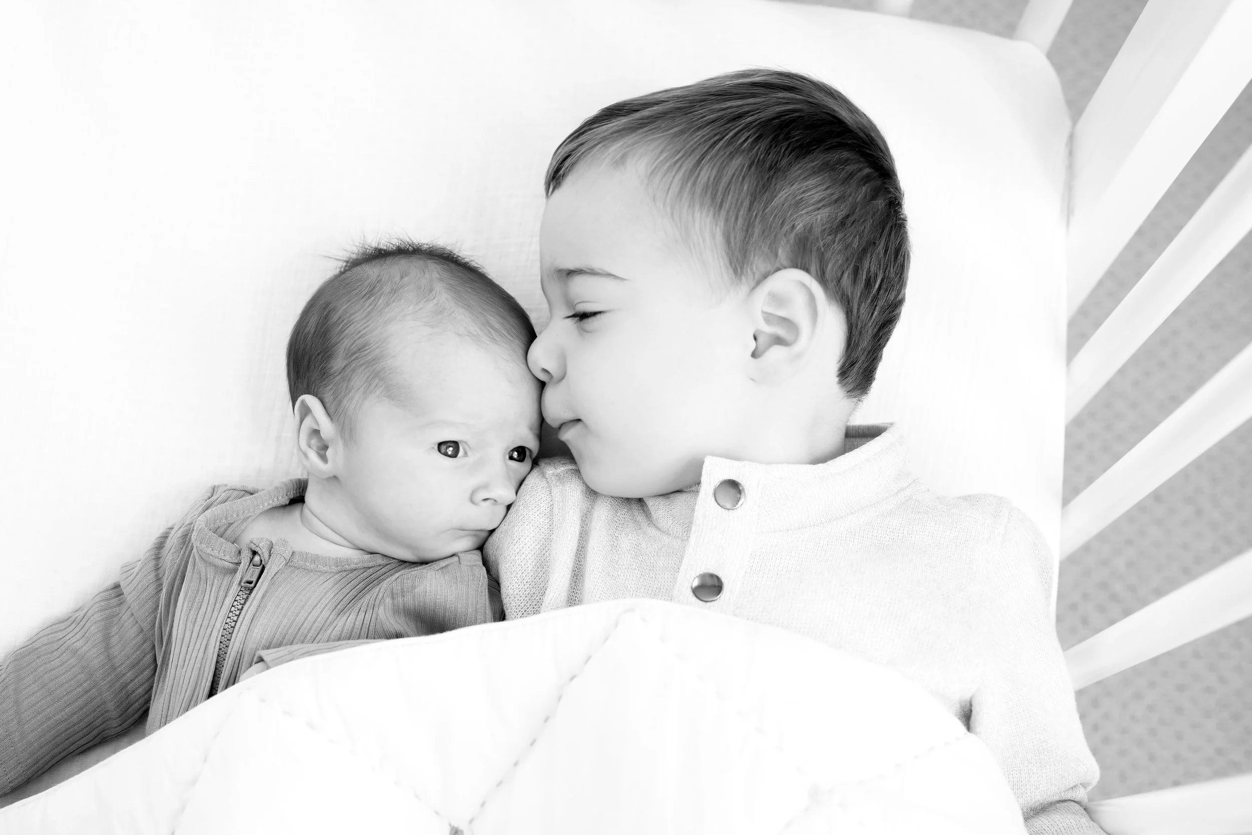 A young boy and a baby, both lying in bed, sharing a moment of affection with the boy gently kissing the baby's forehead.