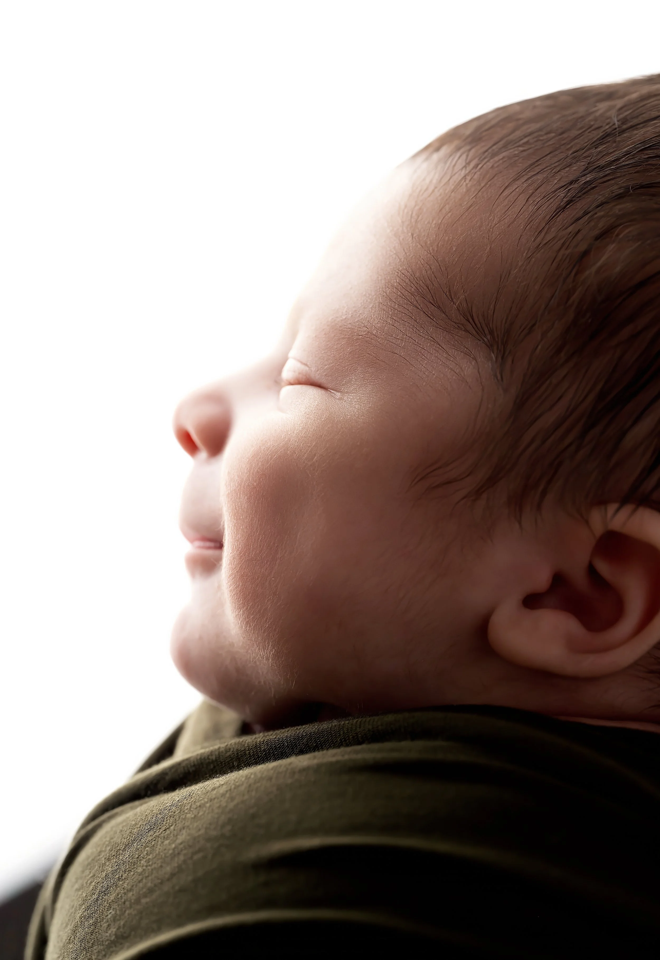 Close-up of a sleeping baby's face in profile, showing closed eyes, nose, and ear, with brown hair and wearing a dark green shirt.