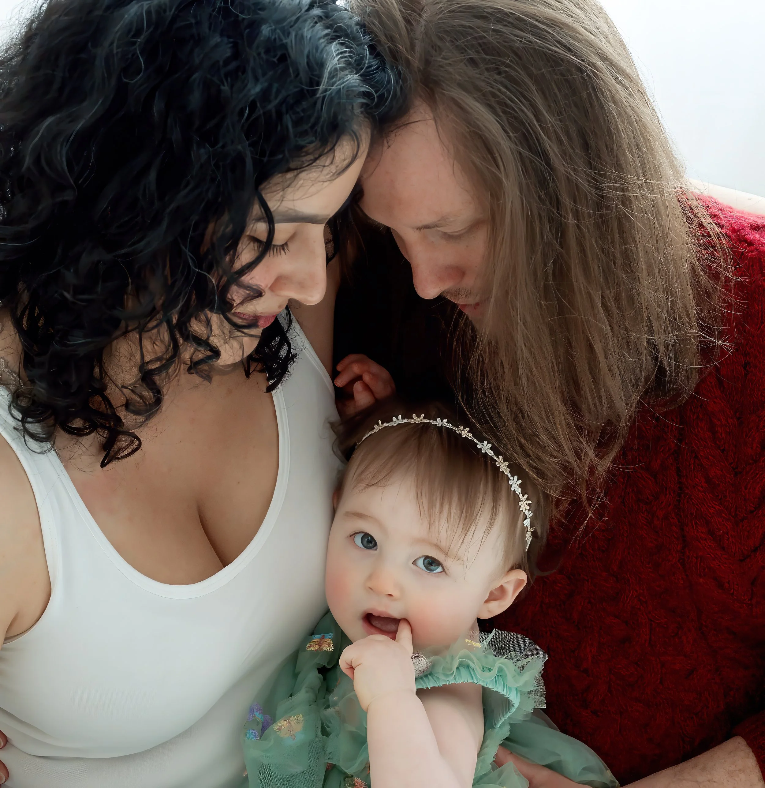 Close-up of a woman with long curly black hair and a young girl with straight light brown hair and blue eyes, laying down on a soft surface with a red blanket. The woman is wearing a white tank top and the girl is wearing a pastel-colored dress with 