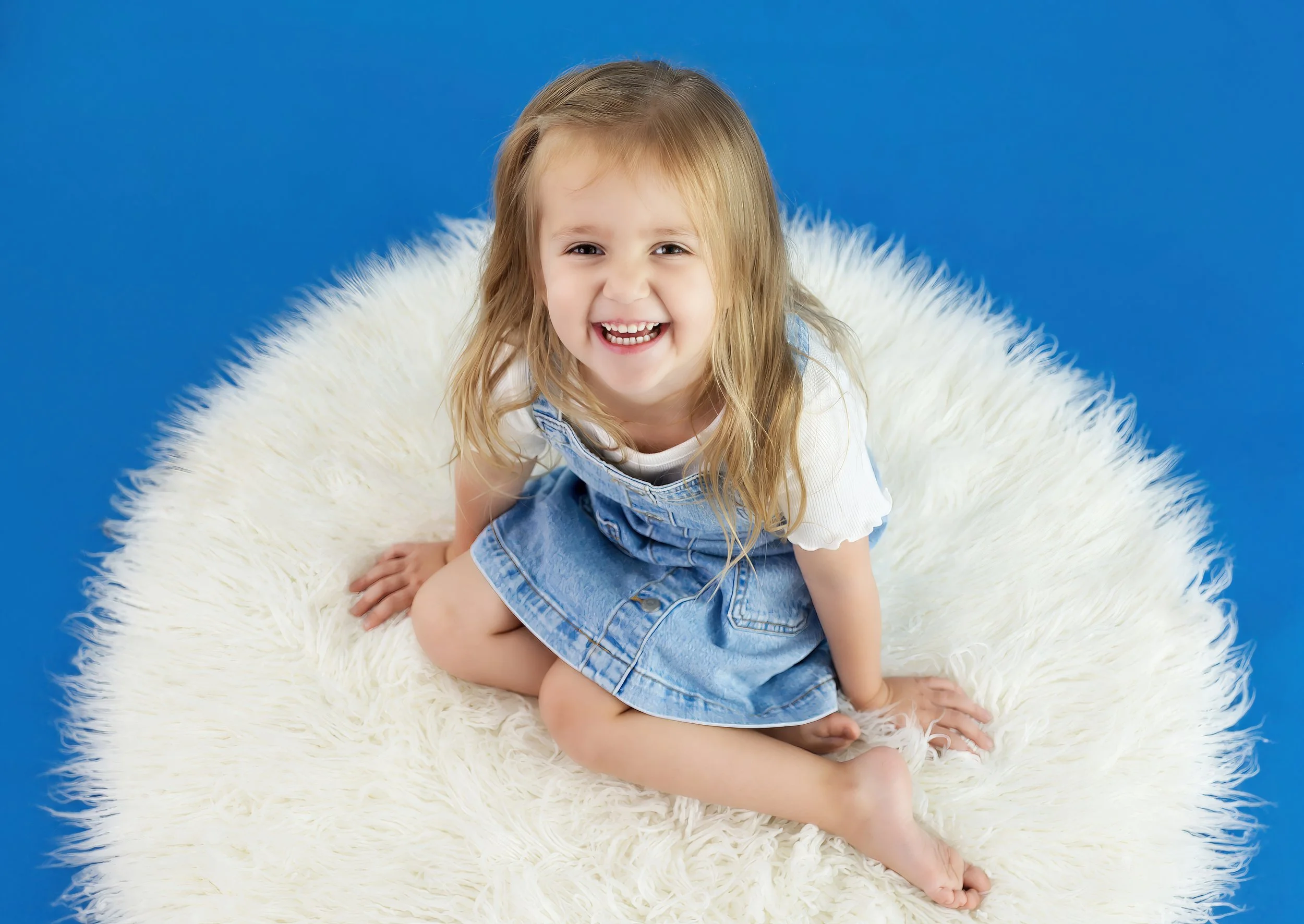 A young girl with long light brown hair smiling, sitting on a white fluffy rug against a blue background, wearing a white shirt and denim overalls.