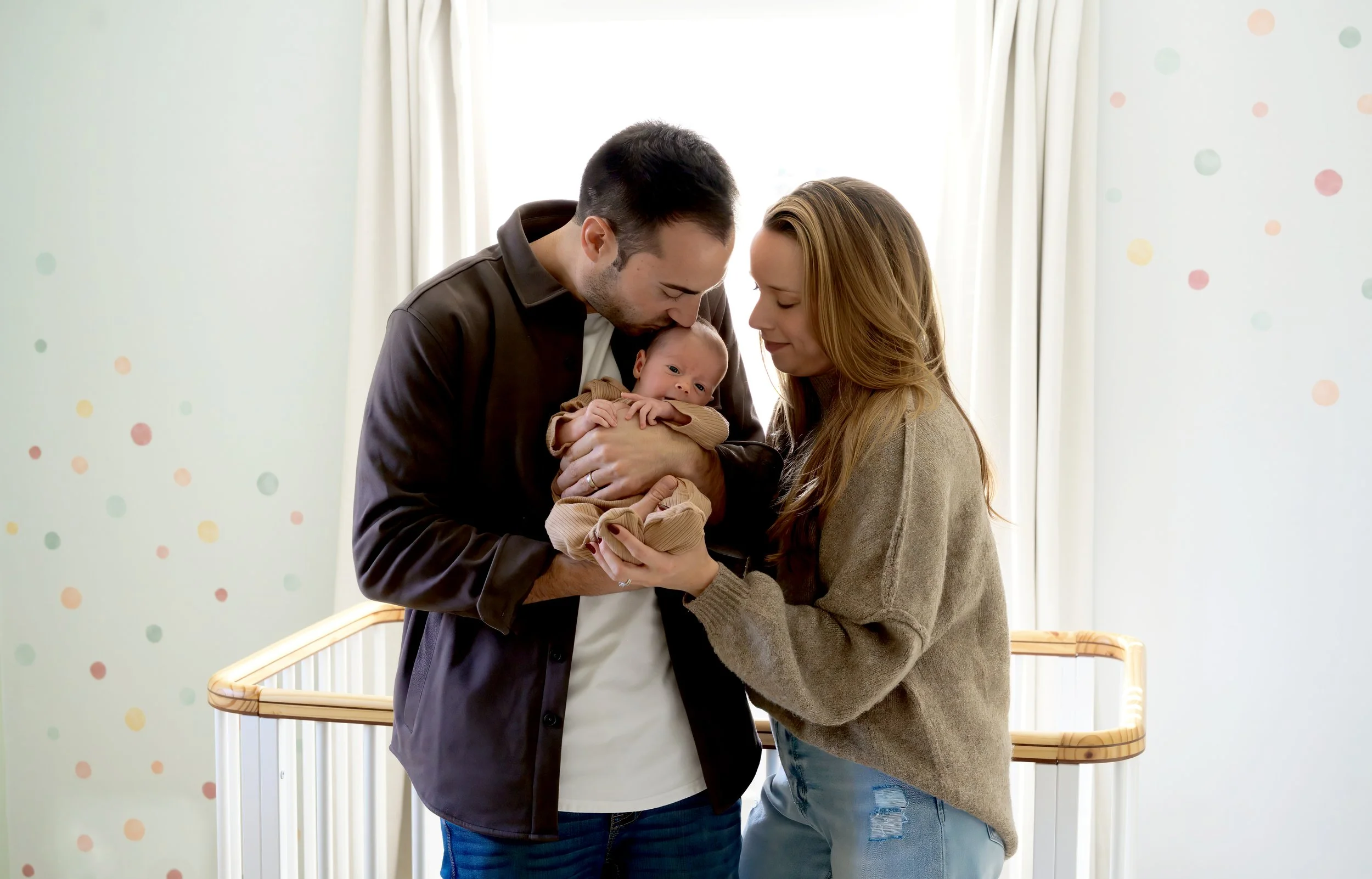 A man and woman holding a newborn baby together in a hospital or maternity unit, standing near a crib, with a bright background and pastel-colored wall dots.