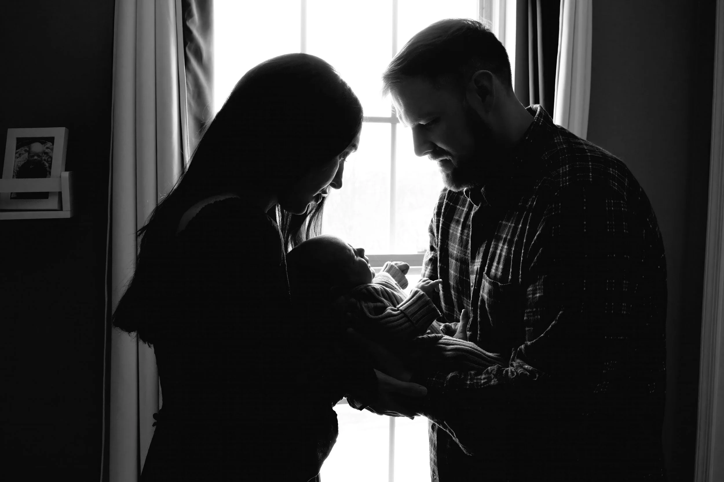 A black and white photo of a man and woman with their foreheads touching, holding a newborn baby in front of a window.