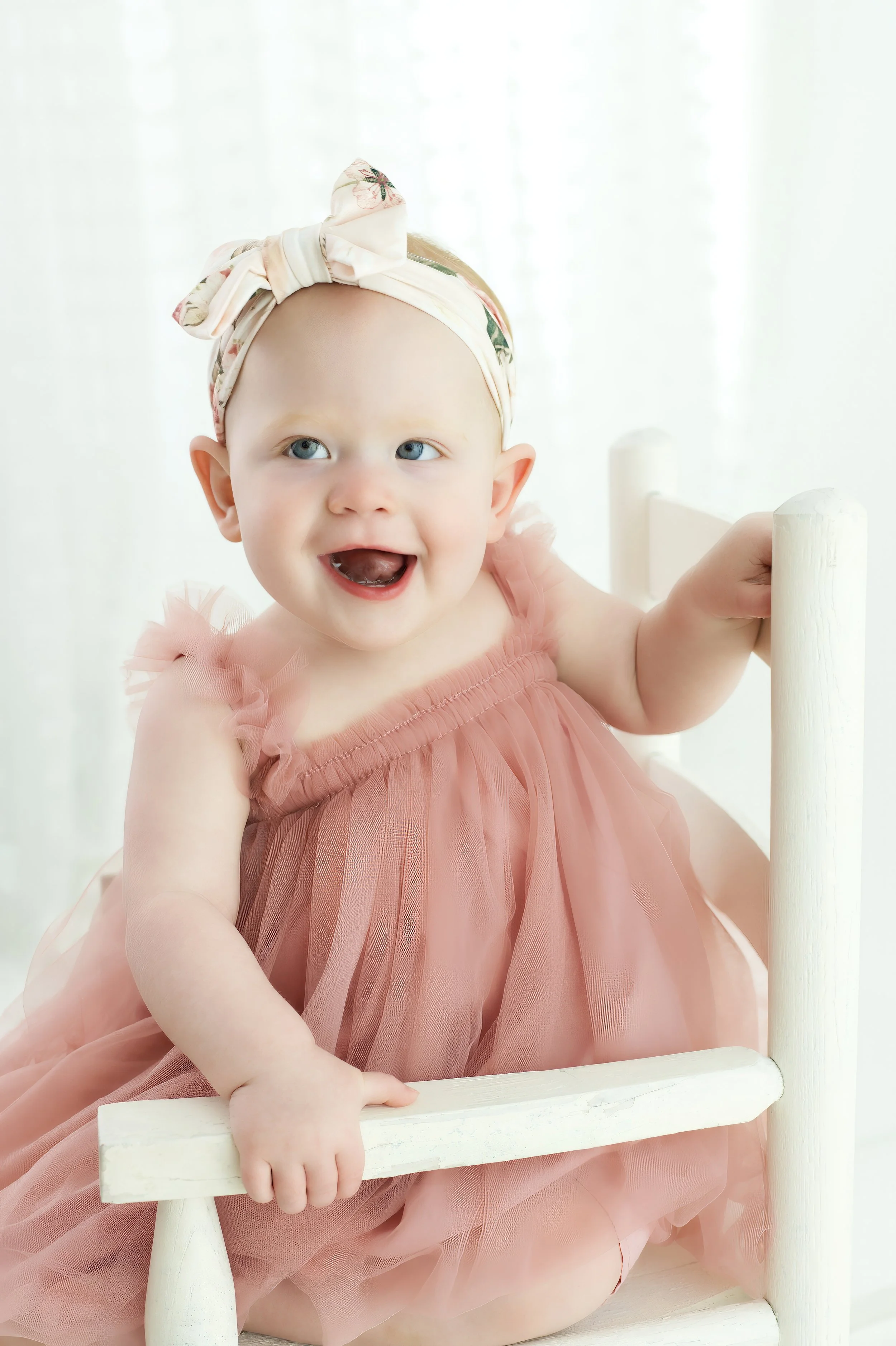 A smiling baby girl with blue eyes wearing a pink tulle dress and a floral headband, sitting on a white chair and holding onto its armrest, with a soft white background.