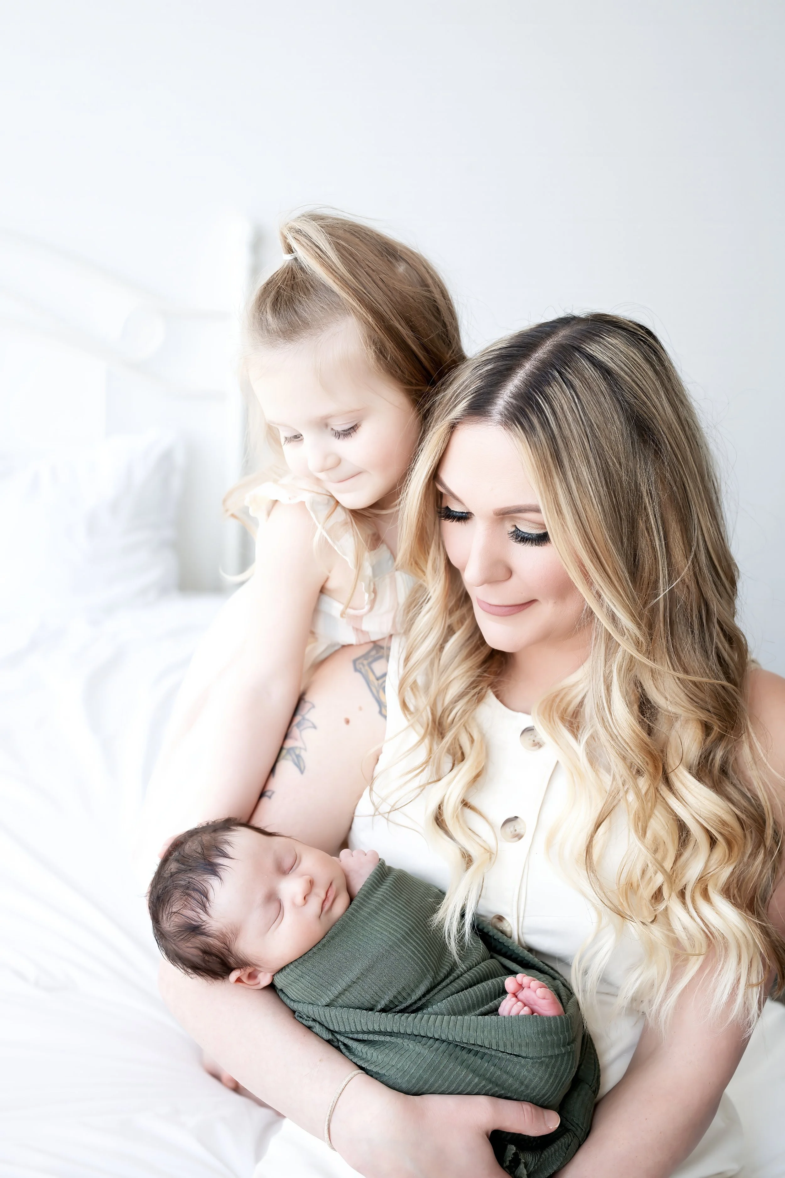 A woman with long, wavy blonde hair holding a sleeping baby in her arms, while a little girl with light brown hair watches and smiles gently. The scene is bright and cozy with white bed linens.