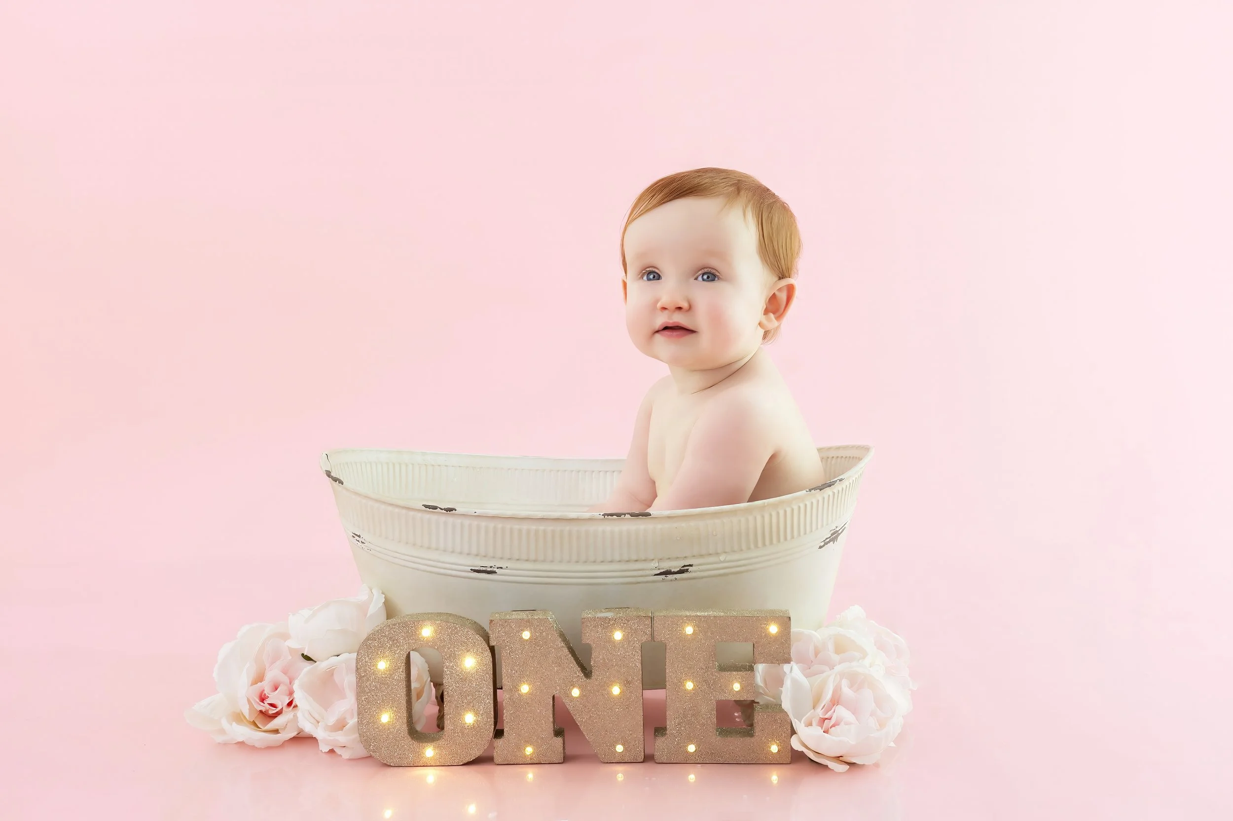 A baby with light skin and red hair sitting in a white tub against a pink background with decorative flowers and a glittery 'ONE' sign.