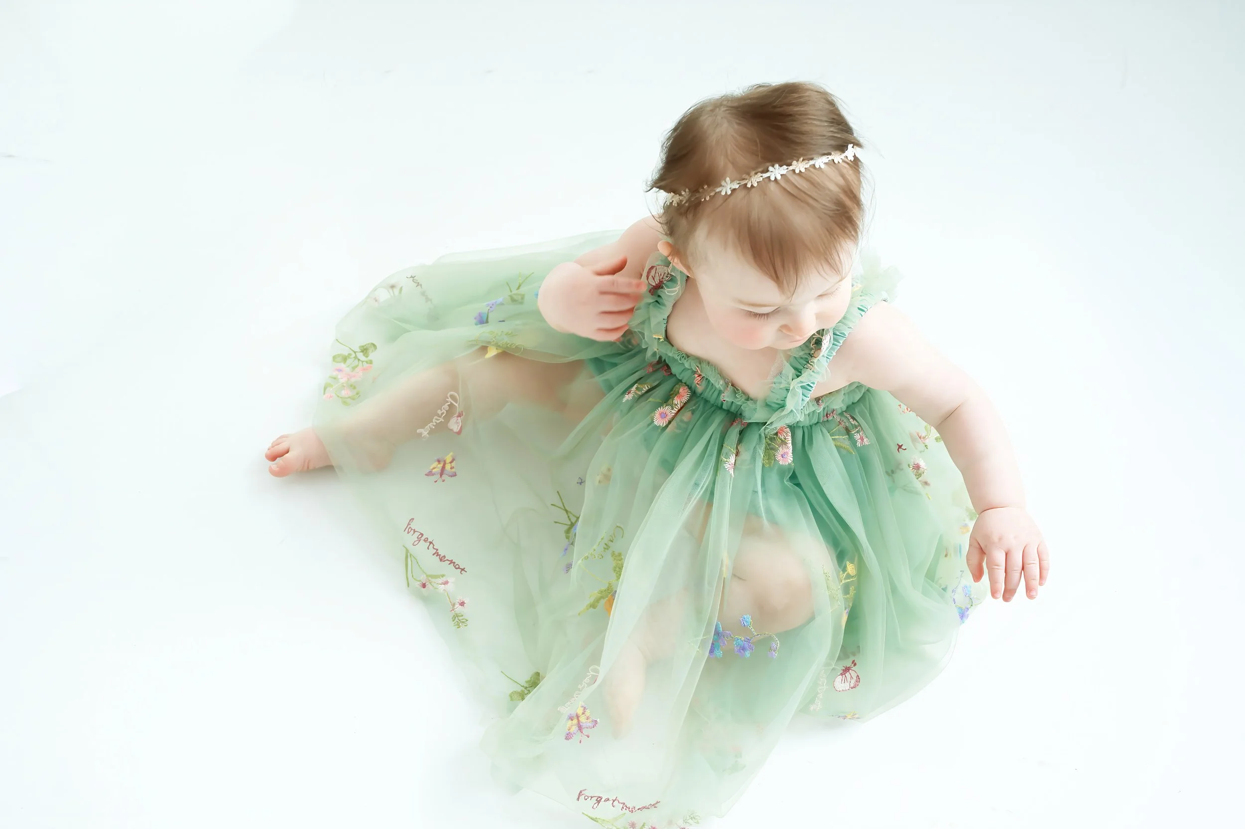 A baby girl with light brown hair wearing a green dress with floral embroidery and a white flower headband, sitting on a white background.