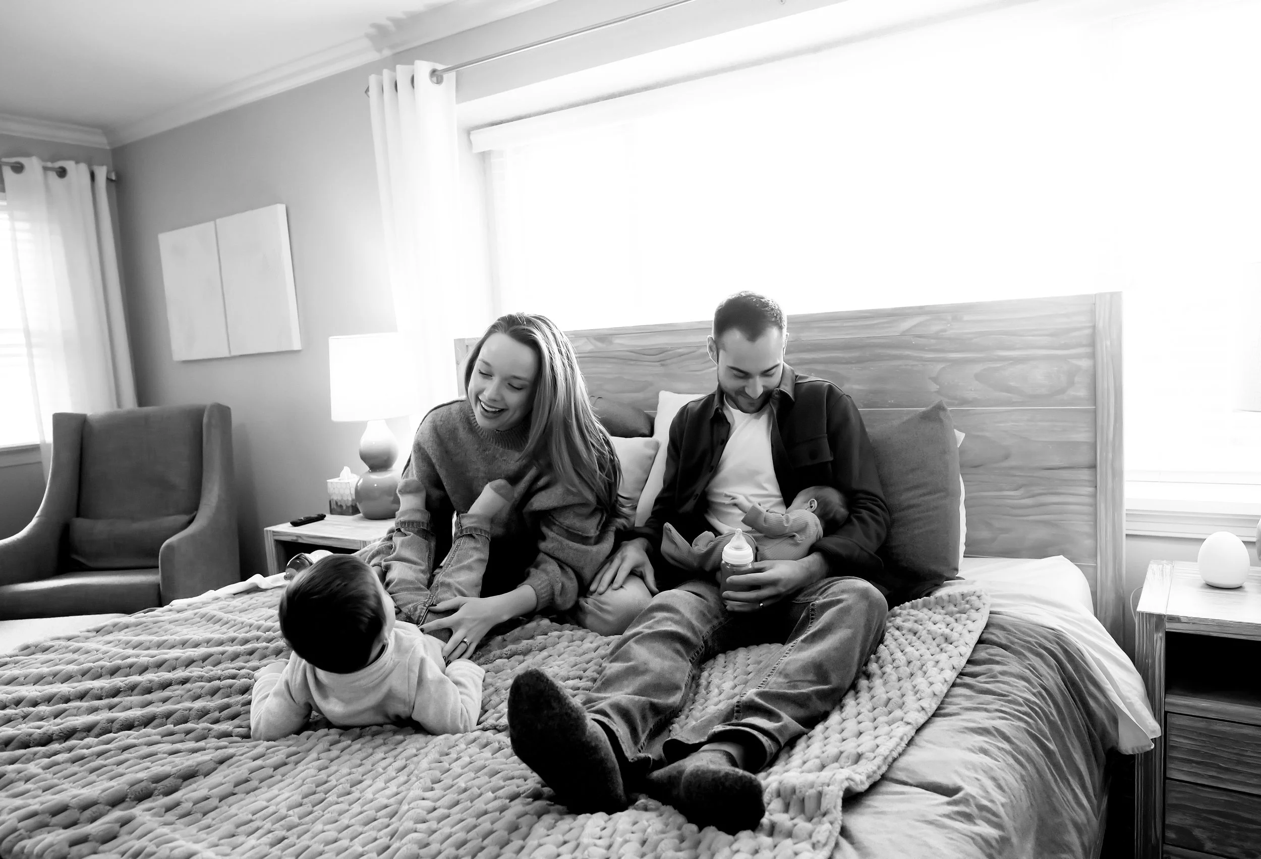 A family sitting on a bed in a bright bedroom, feeding and playing with their young children and baby.
