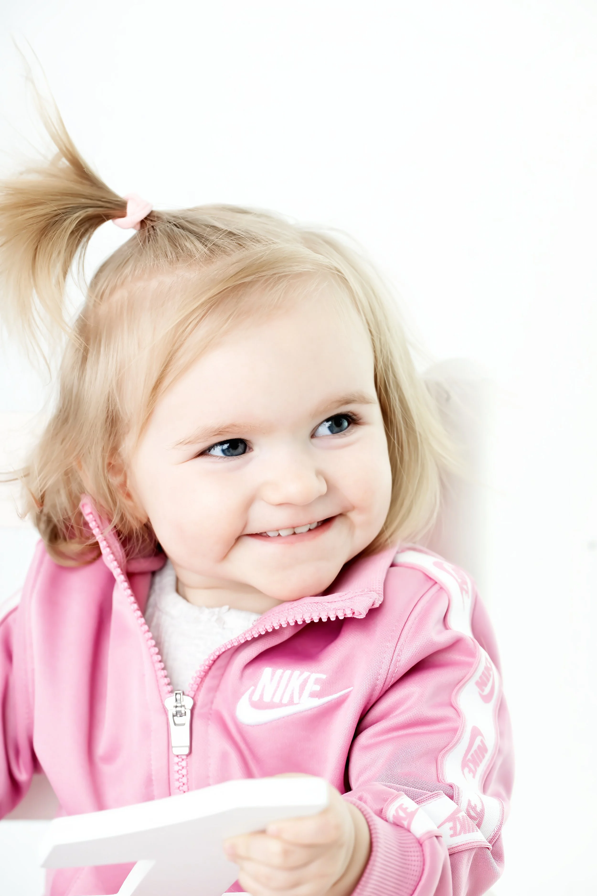 Young girl with blonde hair in pigtails, smiling, wearing a pink Nike jacket, holding a tablet, against a bright white background.