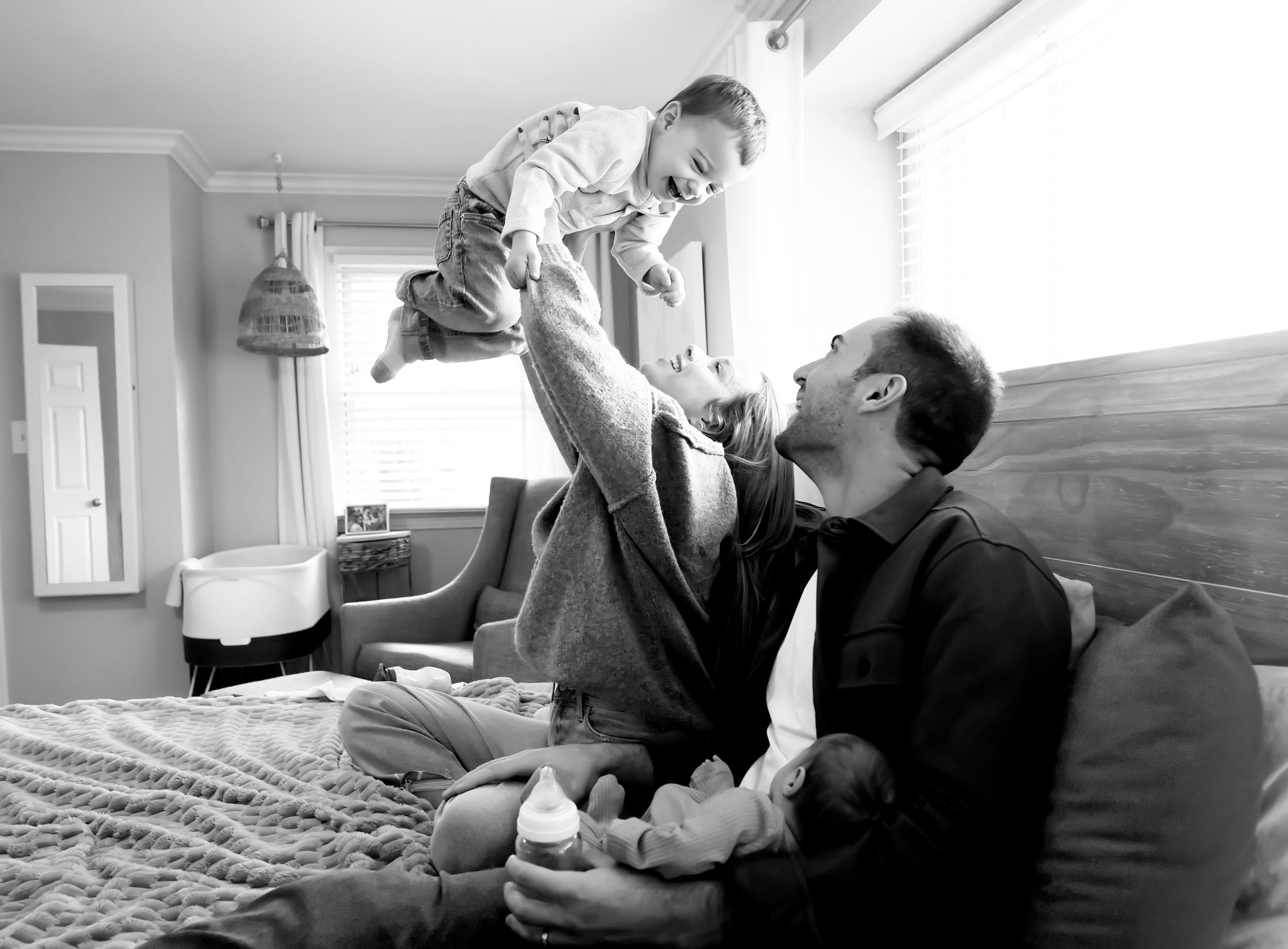 A woman lifting a young boy in the bedroom, both smiling and enjoying playful moment, with a baby lying on the bed.