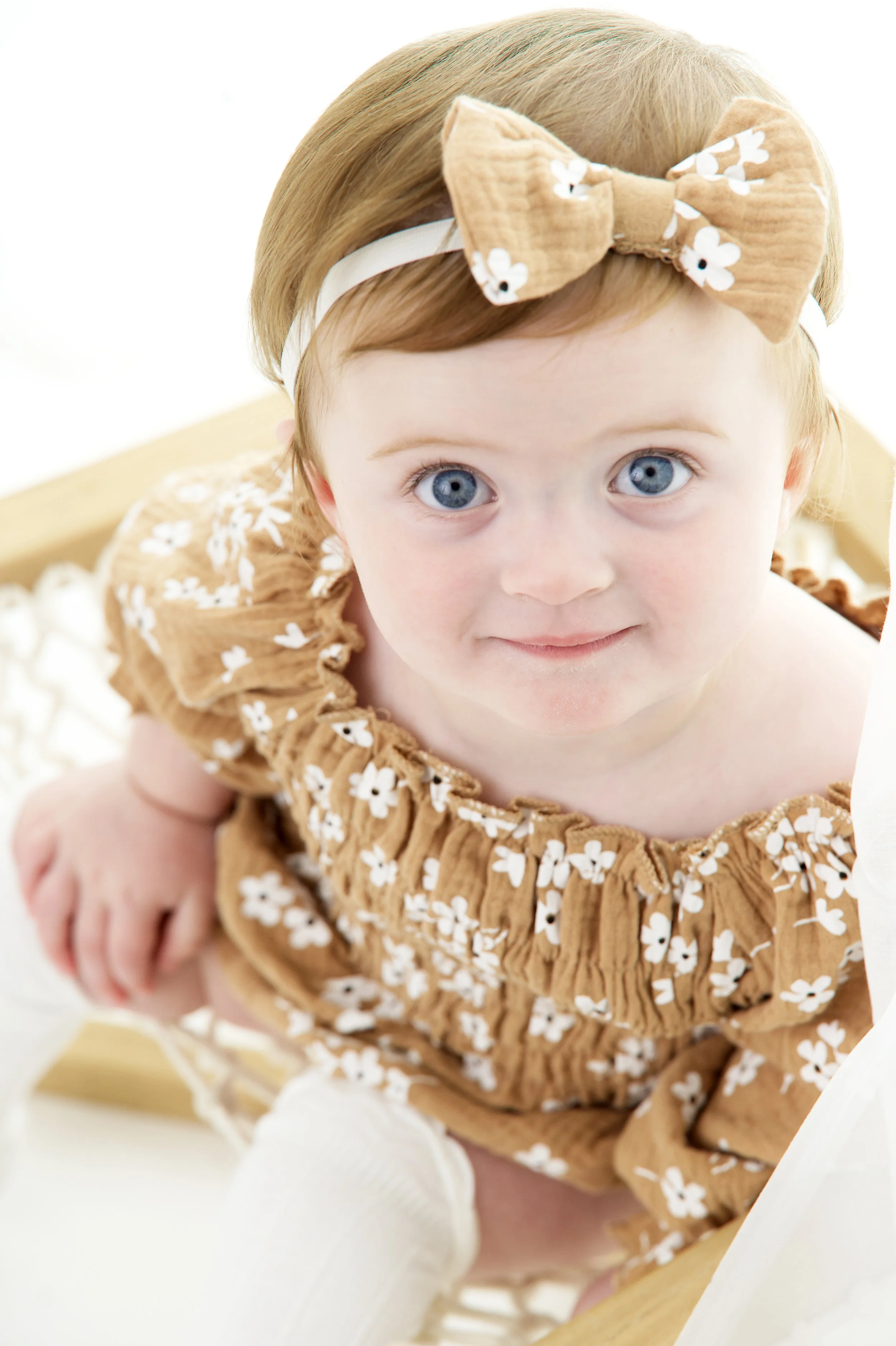 Close-up of a young child with blue eyes, blonde hair, wearing a brown dress with white floral pattern and a matching bow headband, sitting on a wooden high chair, looking at the camera.