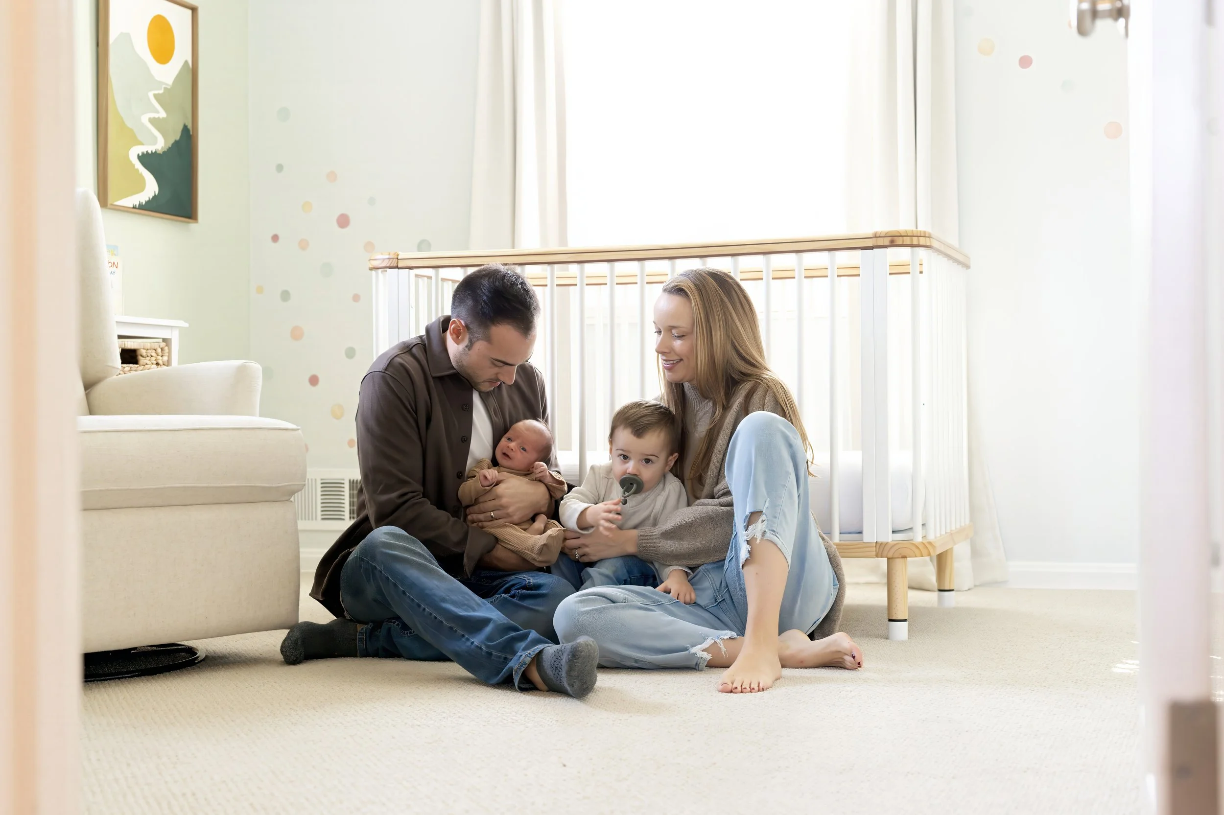 A family sitting on the carpeted floor of a nursery, with a father, mother, and two young children, one of whom is a baby being held by the father. They are smiling and engaging with each other in a bright, cozy room with soft sunlight coming through