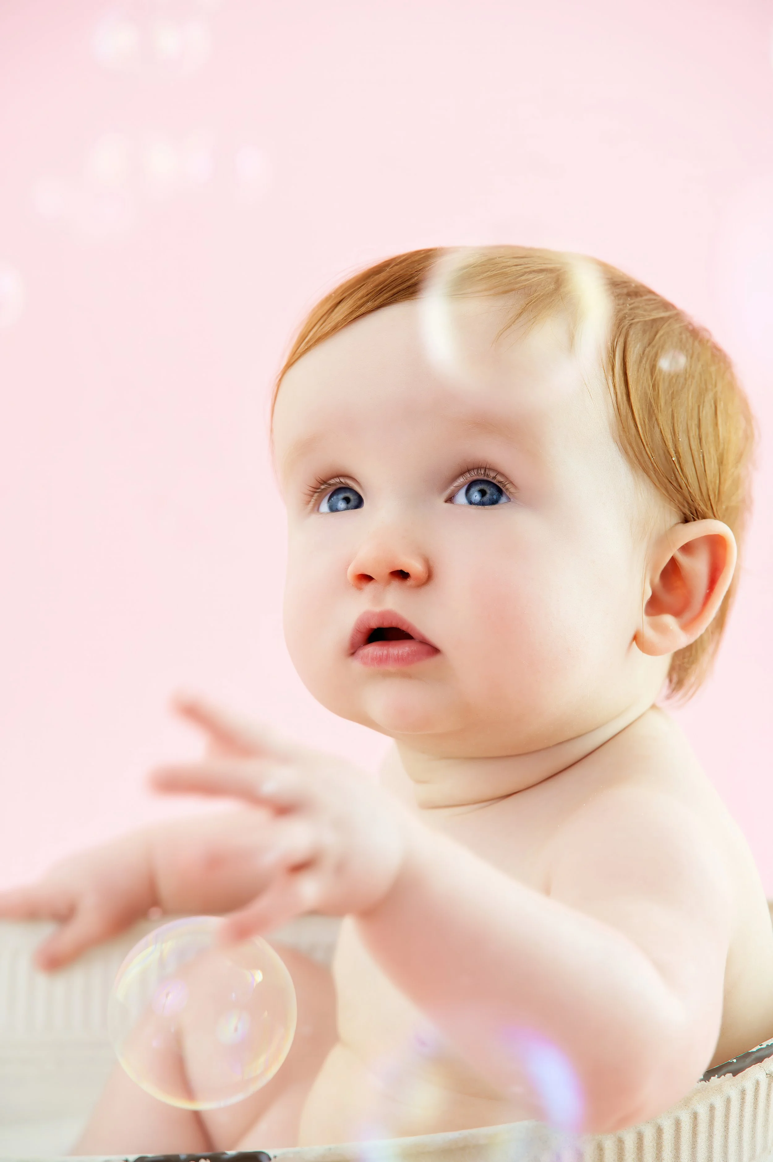 A young child with blue eyes and red hair reaching out towards bubbles against a pink background.
