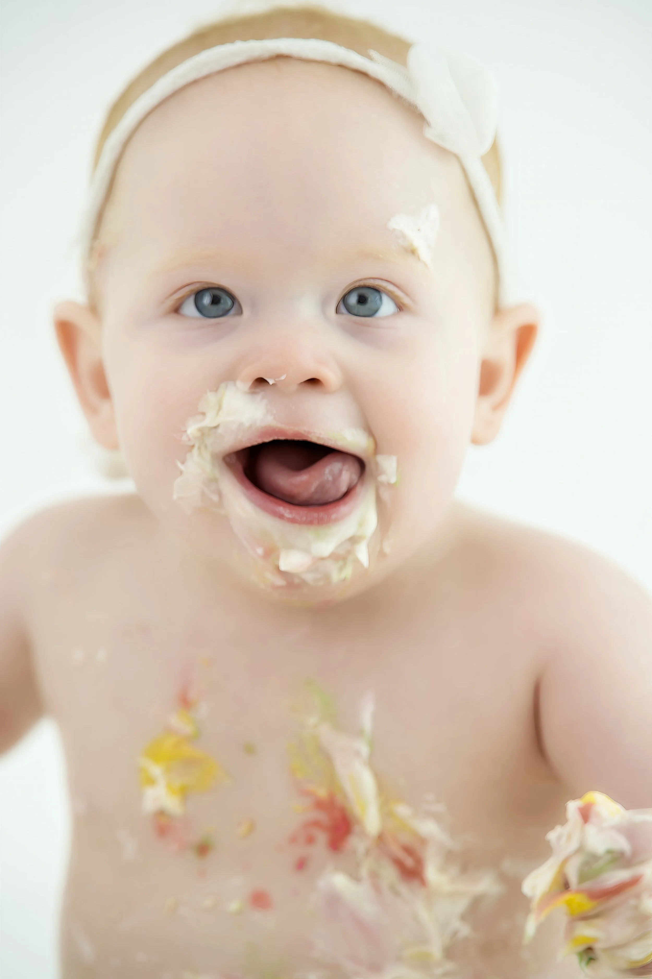 Close-up of a happy baby with blue eyes and a headband, covered in cake and cream, with a bright background.