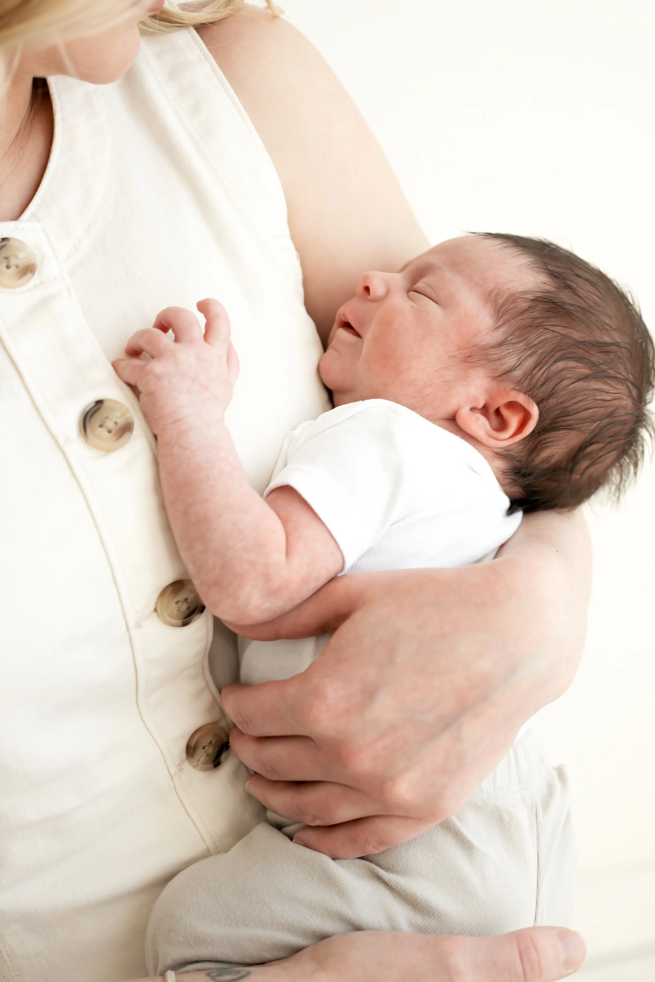 A newborn baby cradled on a woman's chest, smiling with closed eyes, wearing a white outfit, with the woman wearing a cream-colored top with buttons.