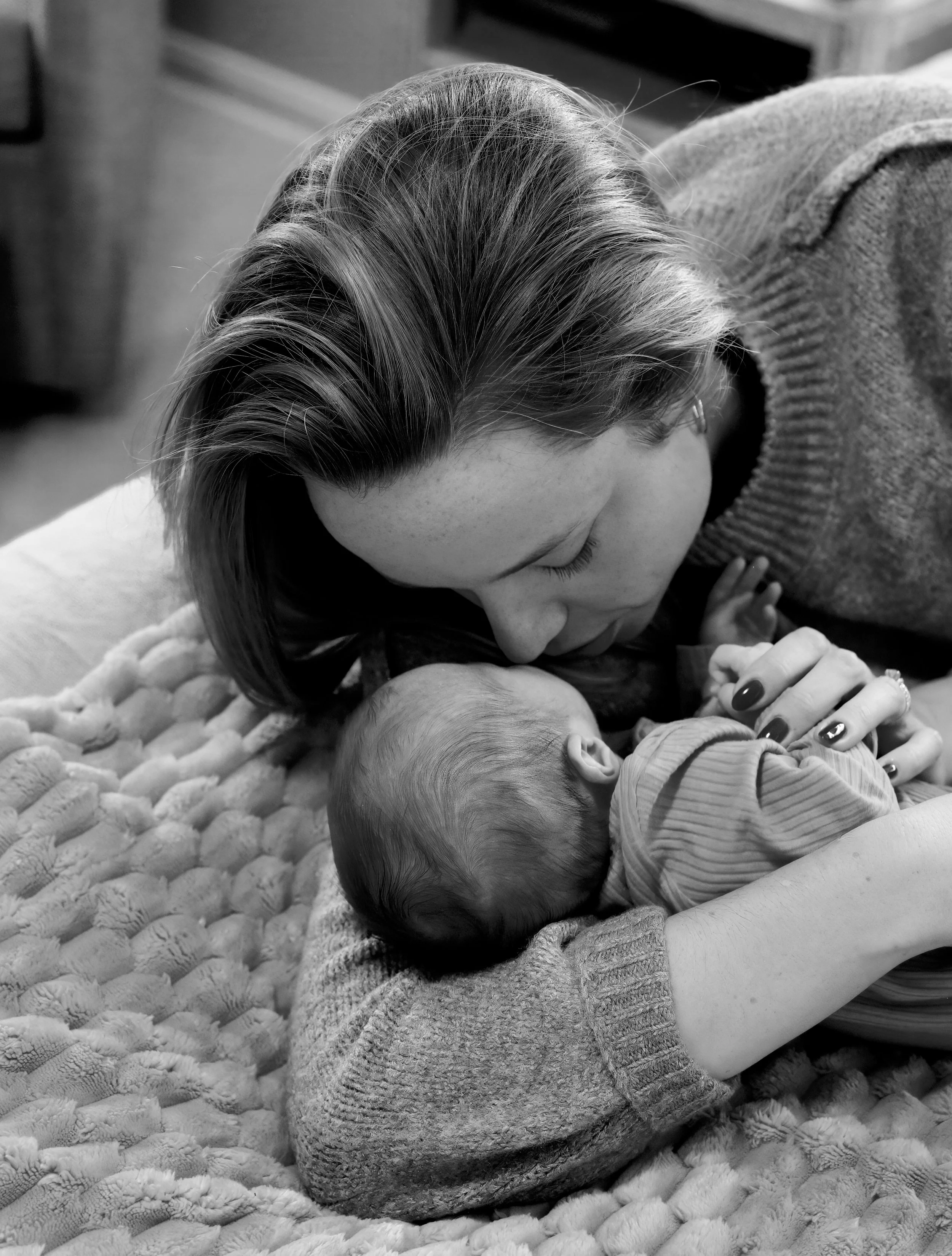 A woman with shoulder-length hair and painted nails is leaning over a sleeping baby on a textured blanket. The woman is wearing a sweater, and the baby has a striped outfit.