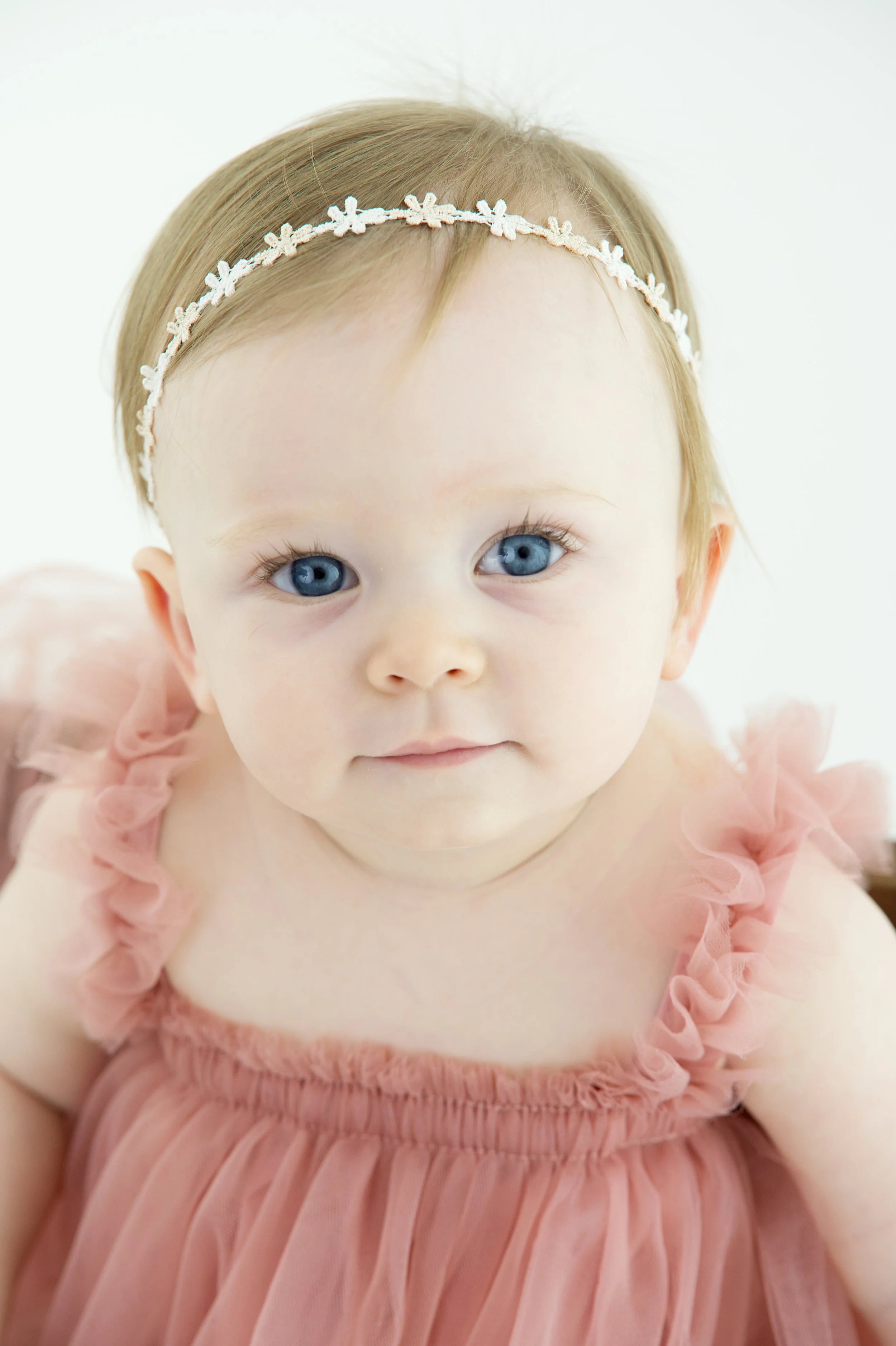 A young girl with blue eyes and blonde hair, wearing a pink ruffled dress and a white floral headband, looking at the camera.