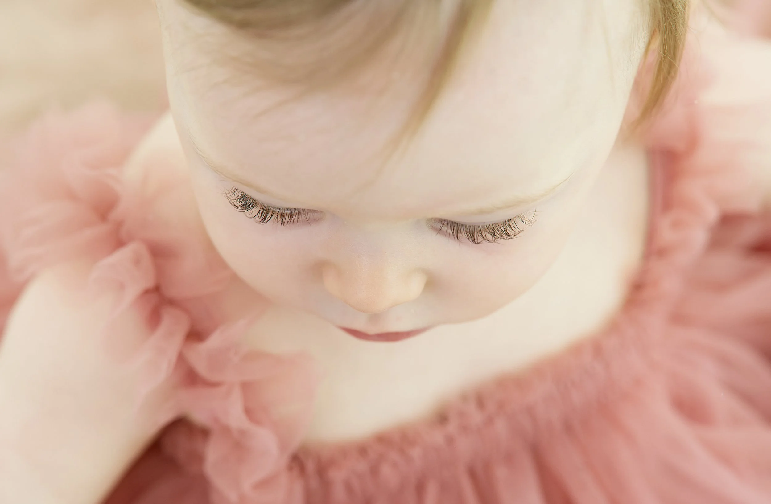 Close-up of a child's face with closed eyes, long eyelashes, and a delicate expression, surrounded by pink fabric like a blanket or clothing.