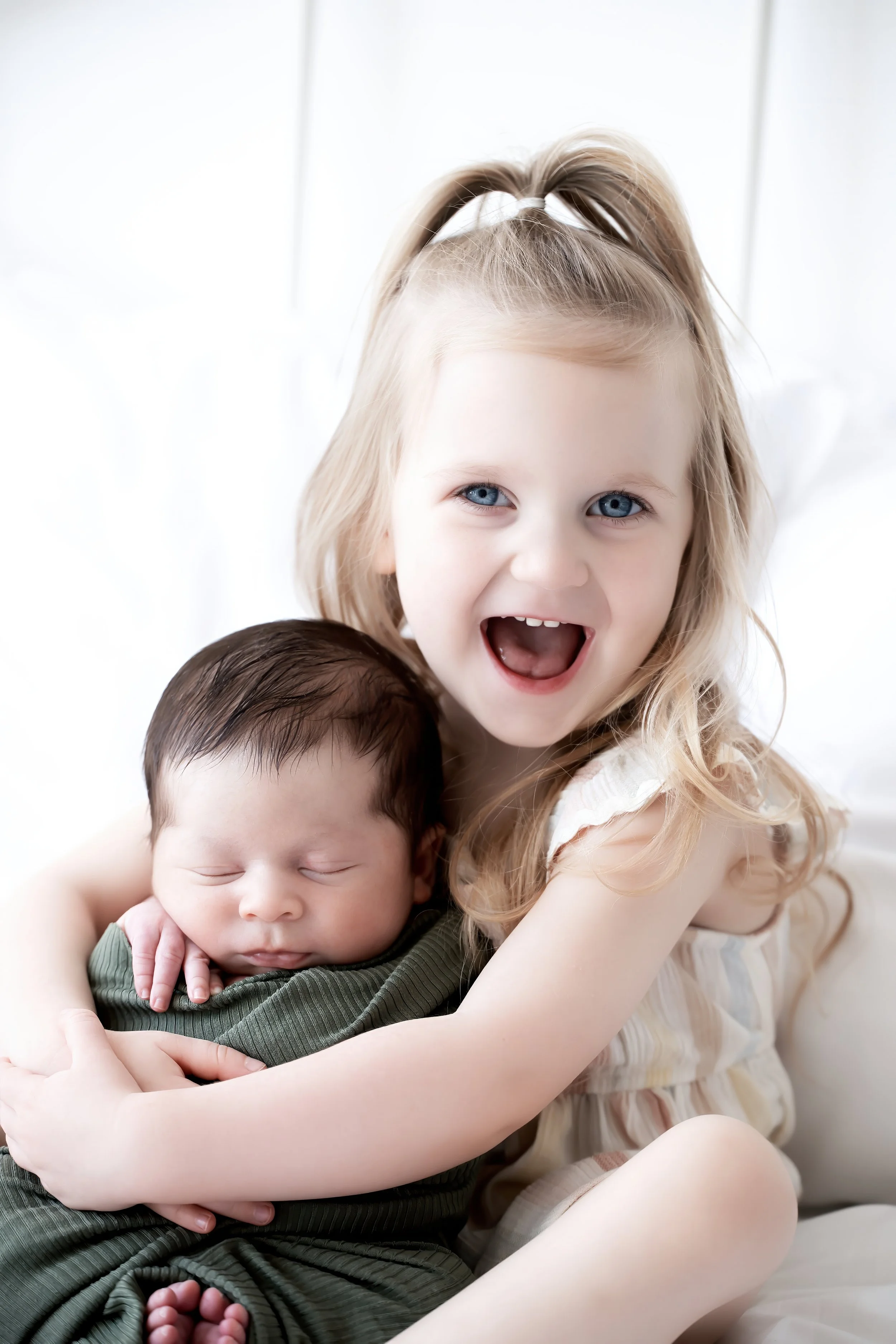 A young girl with blonde hair and blue eyes smiling widely while hugging a sleeping baby boy with dark hair, all against a light background.