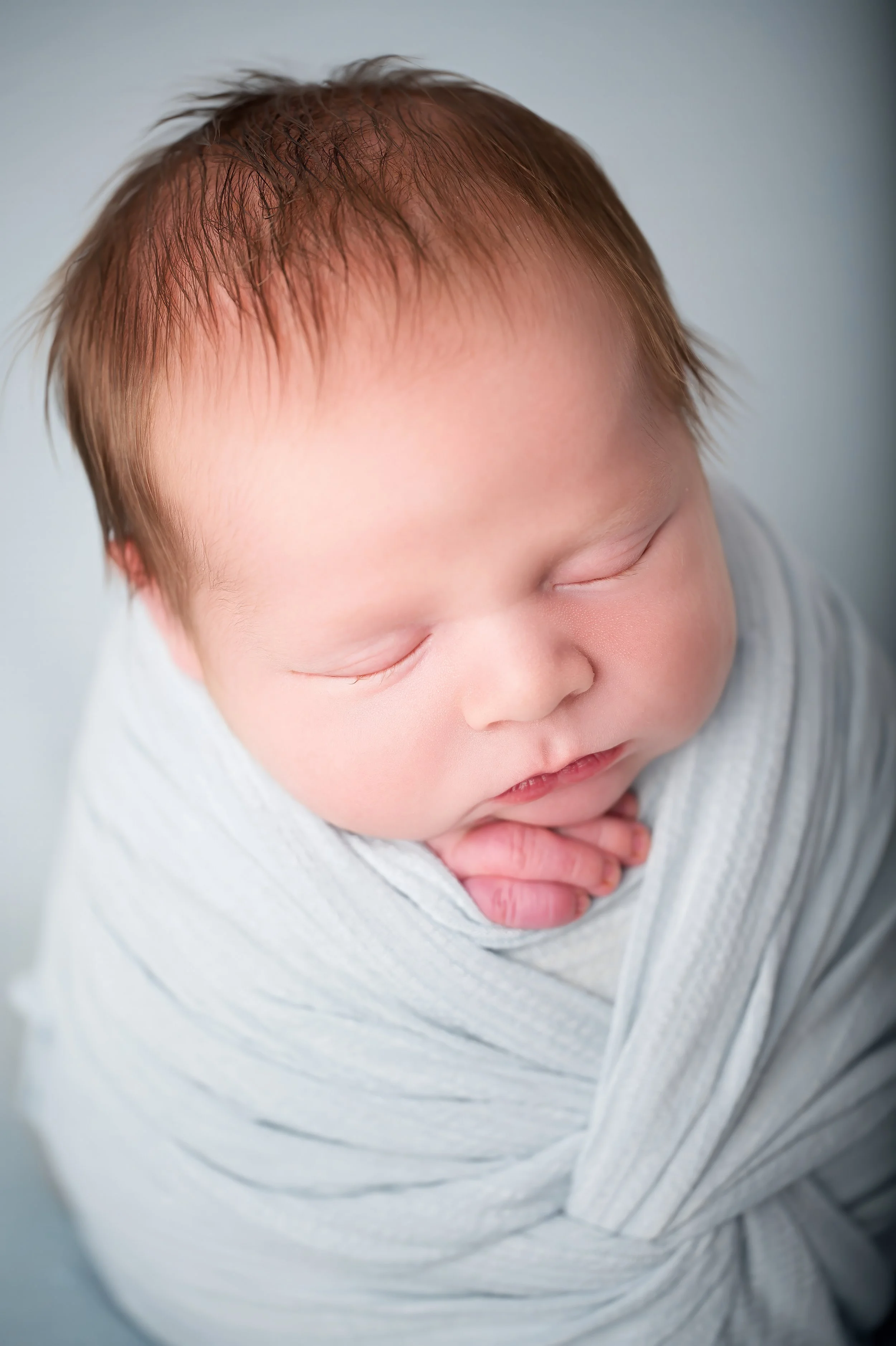 Close-up photo of a newborn baby with brown hair sleeping peacefully, wrapped in a white blanket.