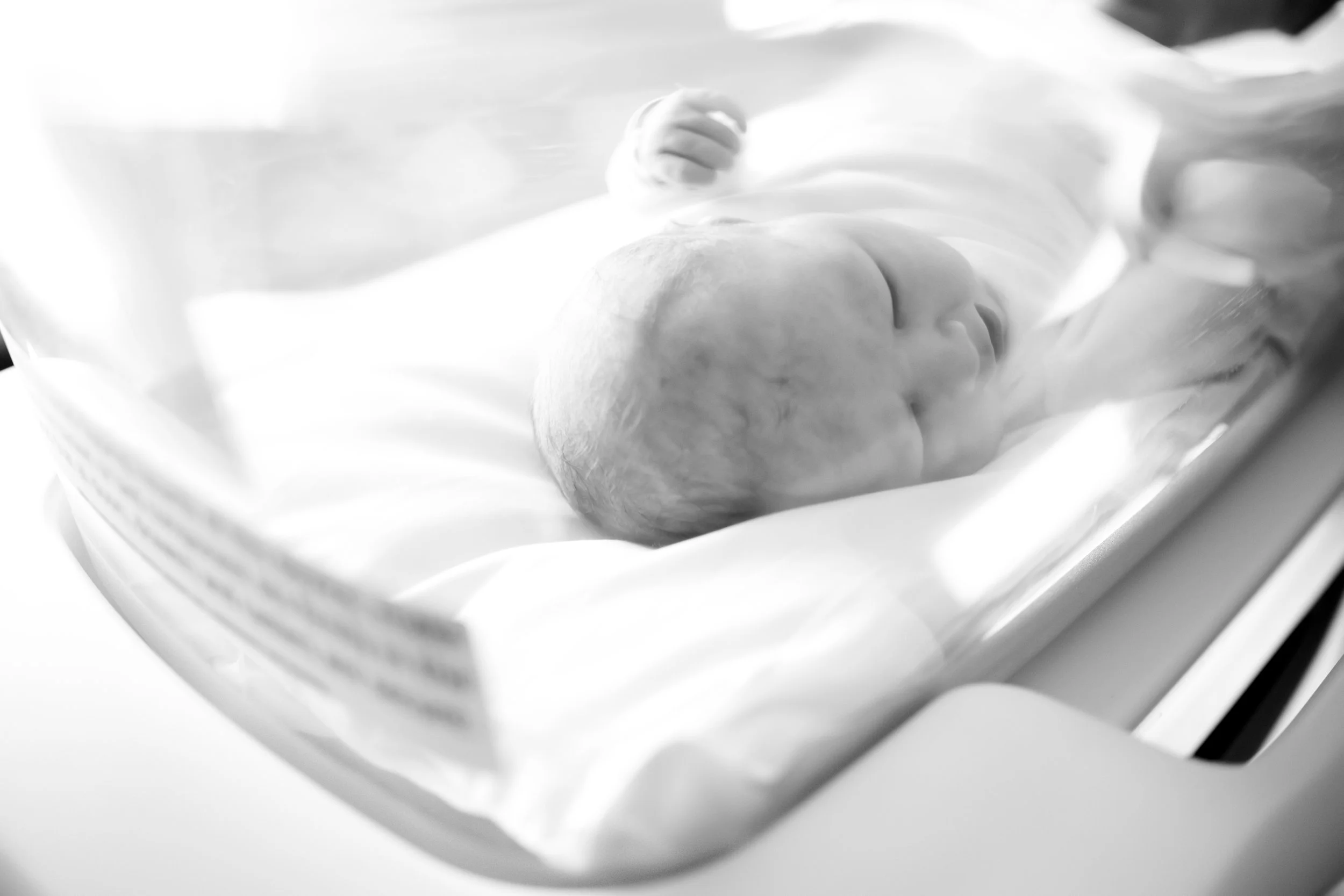 A newborn baby lying on a bed wrapped in a blanket, with a soft focus and grayscale.
