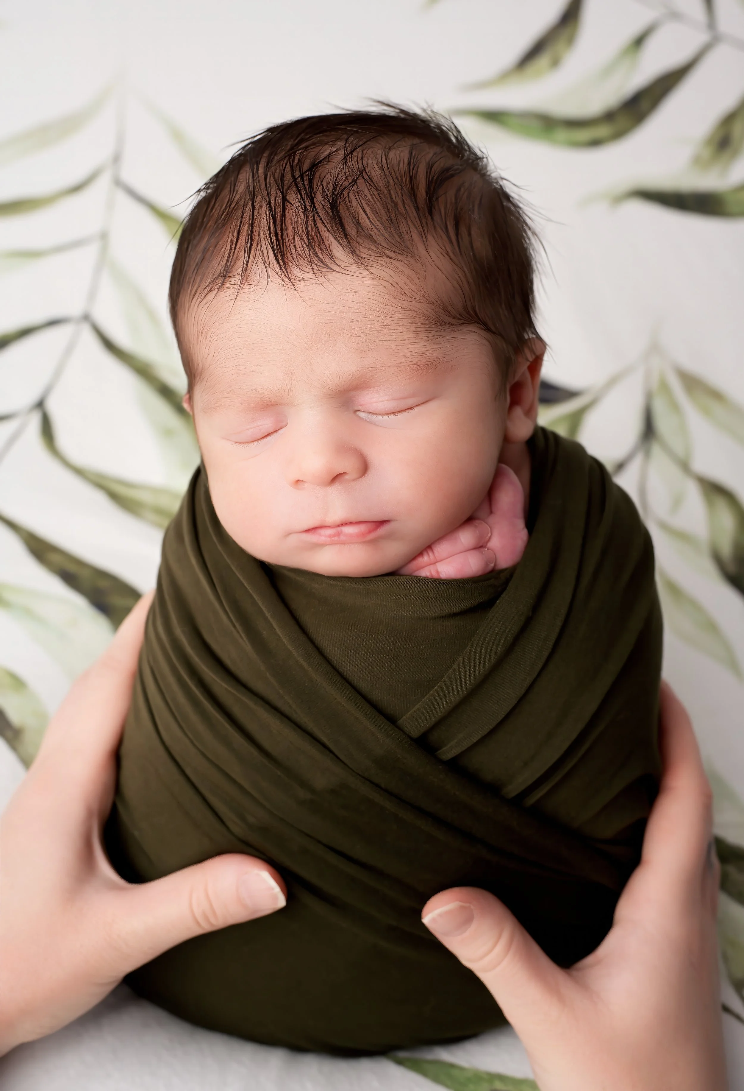 Close-up of a sleeping baby wrapped in dark green fabric, held gently by adult hands, with a leafy background.